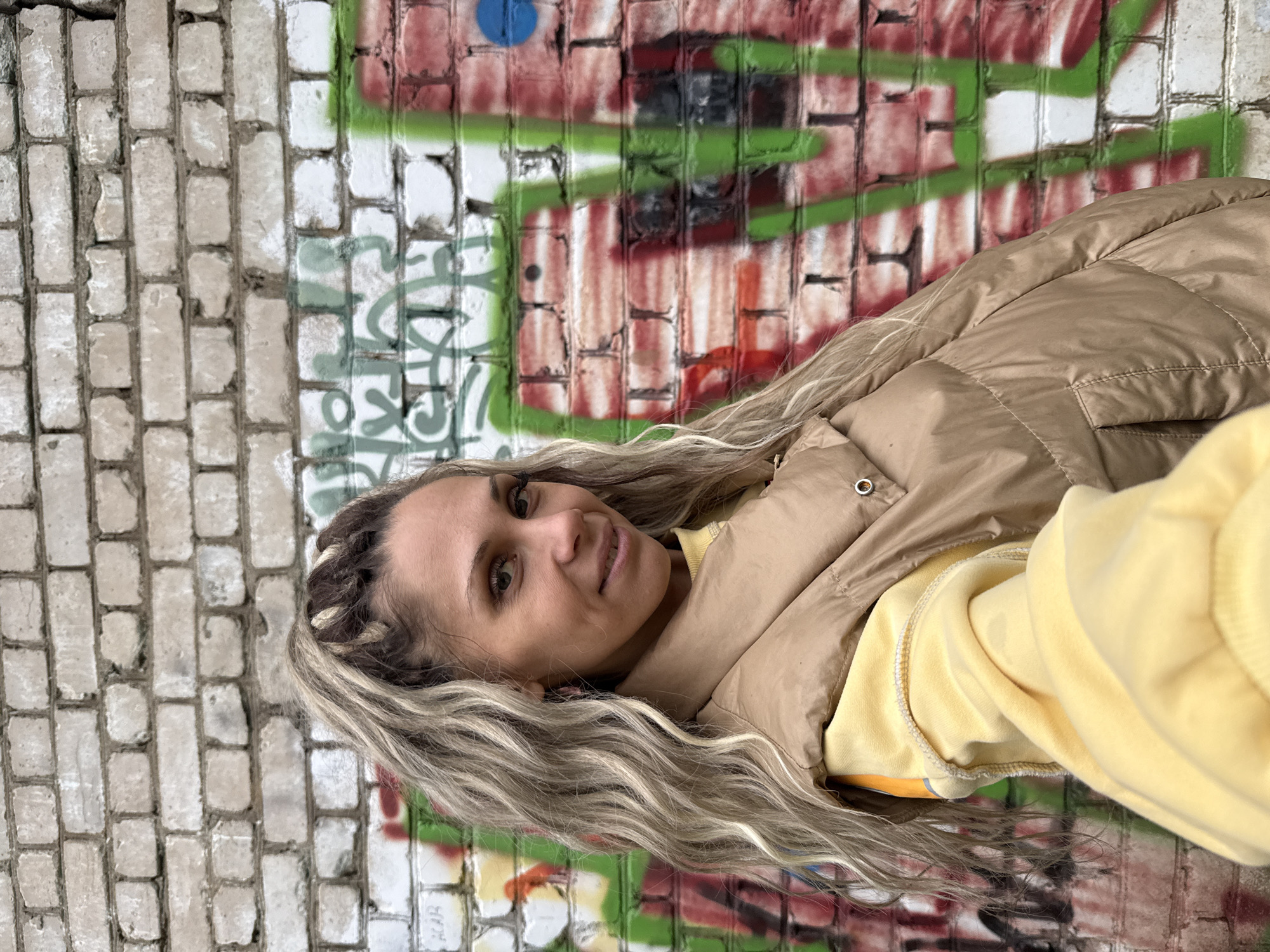 A medium shot captures a woman with blonde, wavy hair styled in a high ponytail with braided accents, standing in front of a graffiti-covered brick wall. She is looking towards the camera with a slight smile. She is wearing a light yellow hoodie and a tan puffer vest over it. The graffiti on the wall features red and green spray paint, with some white brick visible. The image appears to have been taken outdoors, possibly in an urban setting. There is no specific visual information to confirm the location as Rusanovo, Russia.