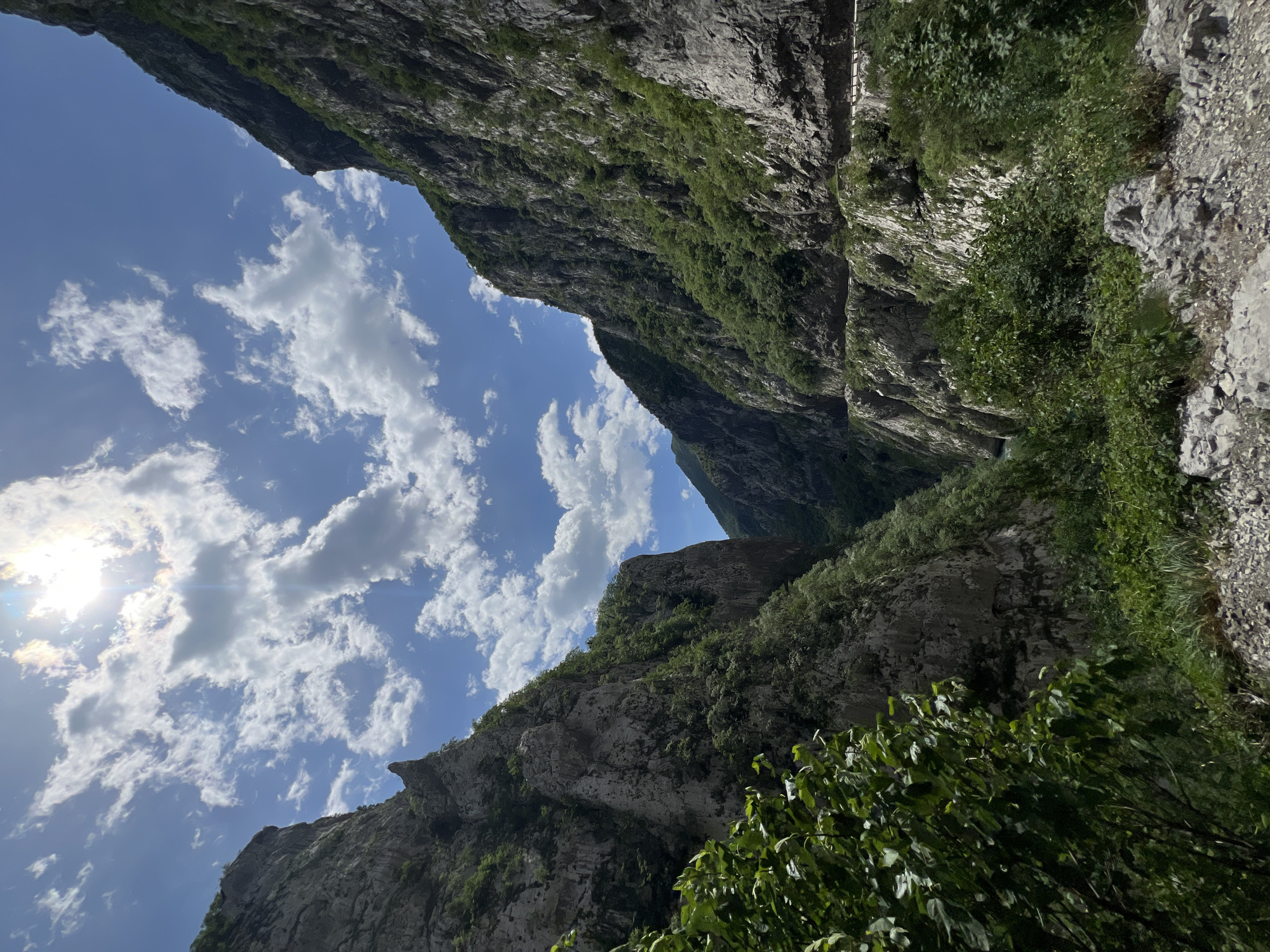 The image shows a deep gorge, likely in a mountainous region. The perspective is looking upwards from the base of the gorge. The sides of the gorge are steep, rocky cliffs covered with some vegetation. The sky is visible between the cliffs, showing blue sky with scattered clouds and a visible sun. The scene is likely in Vlahovići, Montenegro, based on the location provided. There are no people visible in the image. The primary subjects are the geological features of the gorge, the vegetation, and the sky. The environment is natural and rugged.