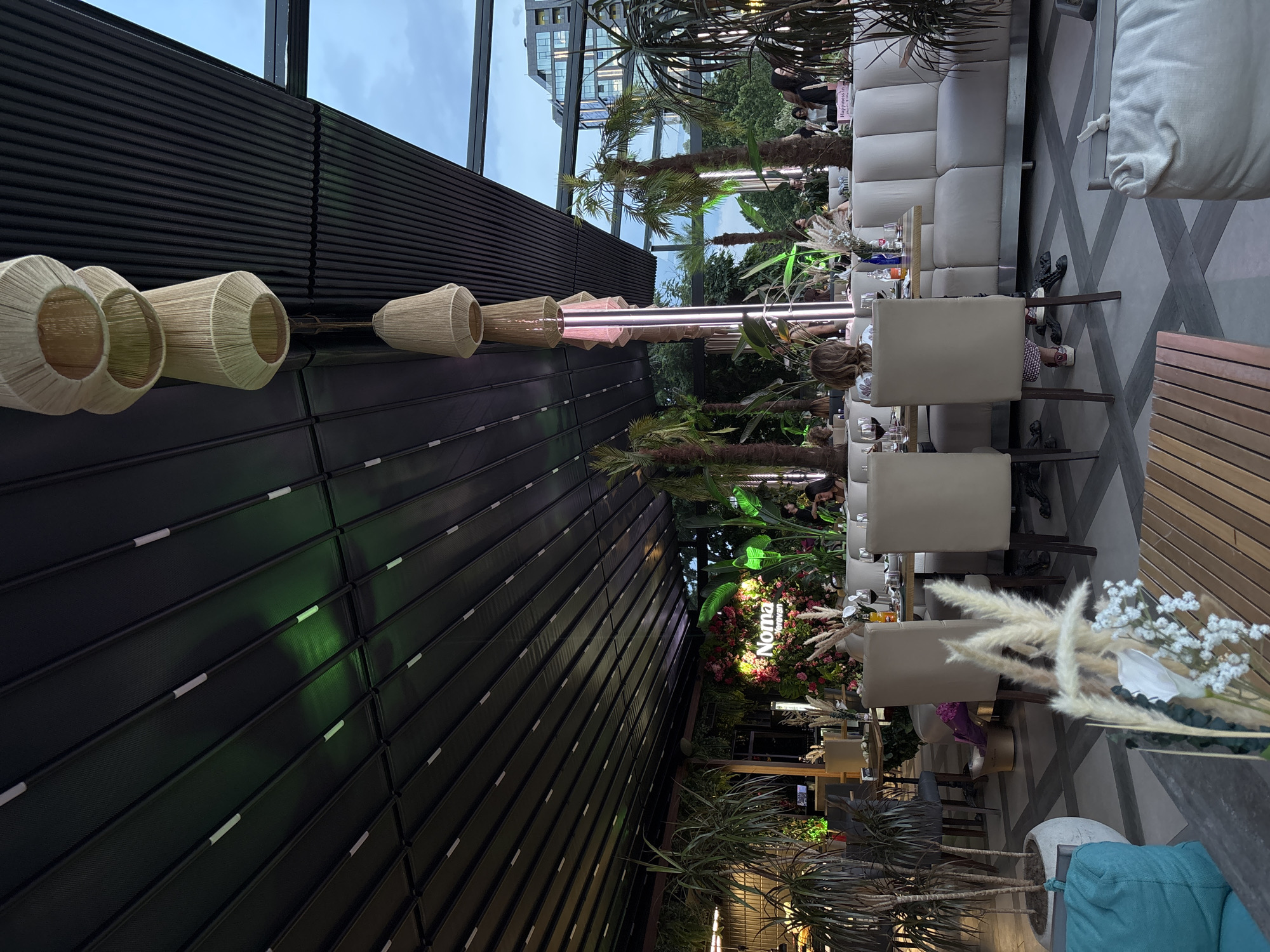 The image depicts an outdoor seating area of a restaurant in Yerevan, Armenia. The scene is set under a retractable roof structure, with a slatted, dark ceiling that has integrated lighting, some of which emits a green glow. Hanging from the ceiling are three large, woven pendant lamps with a neutral, beige tone. The restaurant features lush greenery, including tall potted plants and palm trees, creating a tropical ambiance.
In the foreground, a low wooden table is visible, with a vase containing dried grasses and white flowers. To the left, a large teal cushion is partially in view. Further into the seating area, multiple dining tables are set with chairs and place settings, some with wine glasses. A light-colored, tufted sofa runs along the back of a seating section, with several diners seated at tables adjacent to it. In the middle ground, a floral arrangement with illuminated letters spelling "Noma Yerevan" is prominent, serving as a focal point.
In the background, more diners are visible, and beyond them, trees and buildings suggest an urban setting. The sky is visible through a section of the roof, indicating it is daytime or early evening. The overall atmosphere suggests a modern, stylish dining establishment.