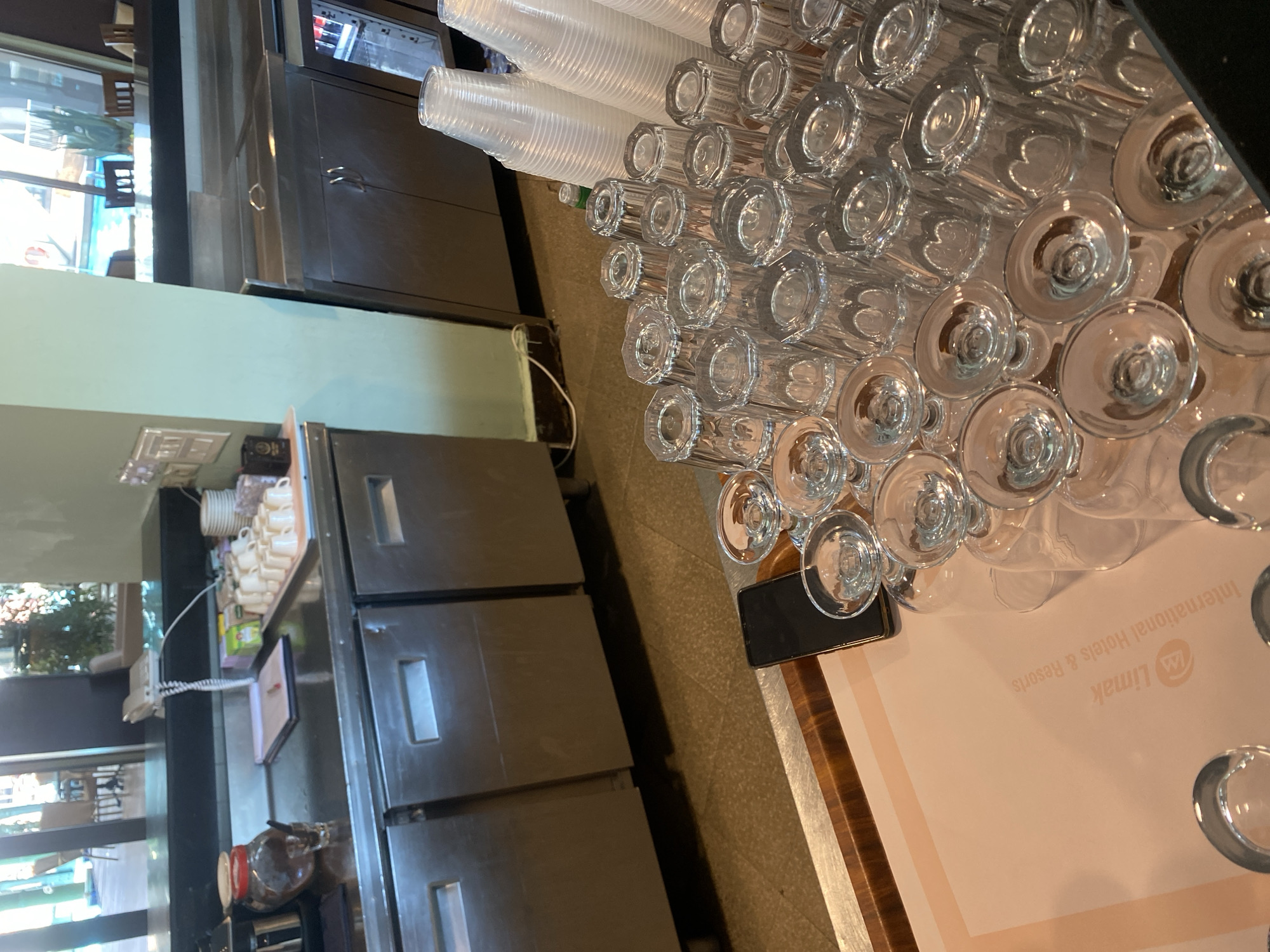 This image depicts a bar or service counter area, likely in a hotel or resort setting, given the text on the tray which reads "Limak International Hotels & Resorts." The foreground is dominated by an array of stacked clear drinking glasses, some inverted and others upright. To the right, several stacks of clear plastic cups are visible. In the mid-ground, a black smartphone rests on a wooden tray. Further back, a stainless steel counter features a tray of white ceramic cups, a jar of coffee beans, a notebook with a pen, and a white telephone. Behind the counter, stainless steel cabinets are visible, and to the far right, a refrigerator unit with drinks inside can be seen. The background shows a glimpse of an outdoor area with tables and chairs, possibly a patio or pool area, illuminated by natural light. The overall impression is one of a functional service area, possibly in the city of Aksu, Turkey, a known location for Limak hotels. There are no people visible in the image.