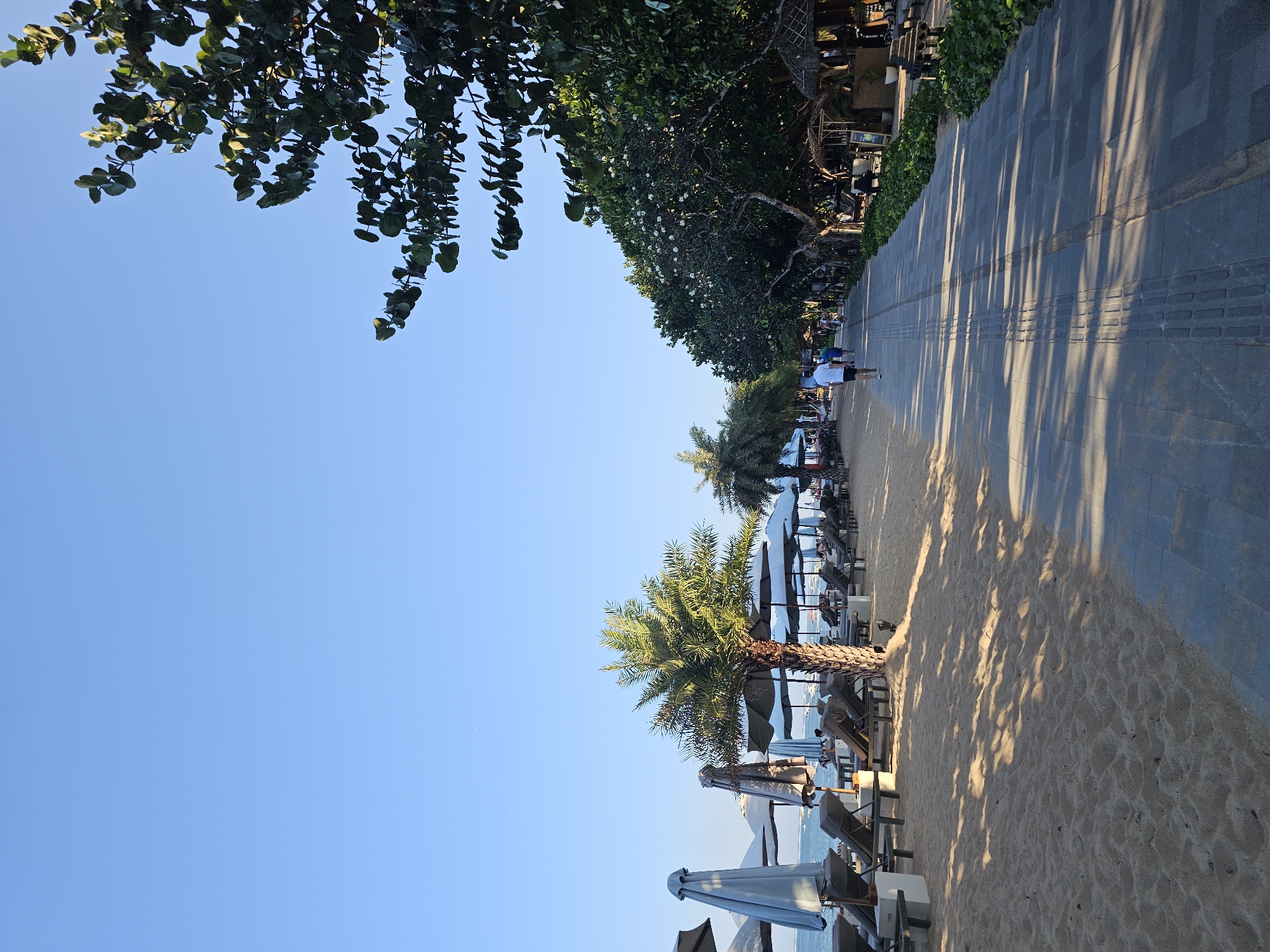 A paved pathway runs alongside a sandy beach in Denpasar, Indonesia. The pathway is bordered on the left by rows of empty lounge chairs and umbrellas, and on the right by lush green trees and vegetation, with some thatched-roof structures visible. A few people are walking along the pathway, some appearing to be tourists. The ocean is visible in the background, with boats anchored offshore. The sky is clear and blue, indicating a sunny day. The lighting suggests it is either early morning or late afternoon, with long shadows cast across the pathway and sand.