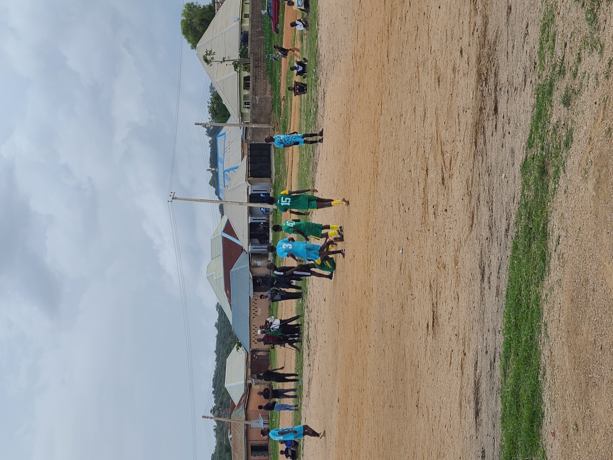 The image depicts a dusty, outdoor scene in Jos, Nigeria, likely a sports field, with a group of young men engaged in a game of soccer. The ground is predominantly sand and dirt, with patches of sparse green grass. Several buildings, characteristic of residential or local community structures, are visible in the background, suggesting an urban or semi-urban environment. The sky is overcast, indicating a cloudy day.
In the foreground, two young men in blue and green soccer uniforms are actively playing, with one in blue dribbling a ball and the other in green preparing to defend. A third player in a green uniform with the number 15 on his back stands nearby, observing the game. A fourth player in a blue uniform is walking away from the center of the action.
In the mid-ground, a cluster of men and boys are gathered, some watching the game, others standing or sitting. Some are dressed casually, while others appear to be involved in the organization or spectating of the match. Among them, a group of men in dark clothing appear to be observing the game from the sidelines.
The environment is characterized by the presence of utility poles and wires, indicating infrastructure. The overall impression is a community gathering centered around a soccer match in a developing urban area of Jos, Nigeria.