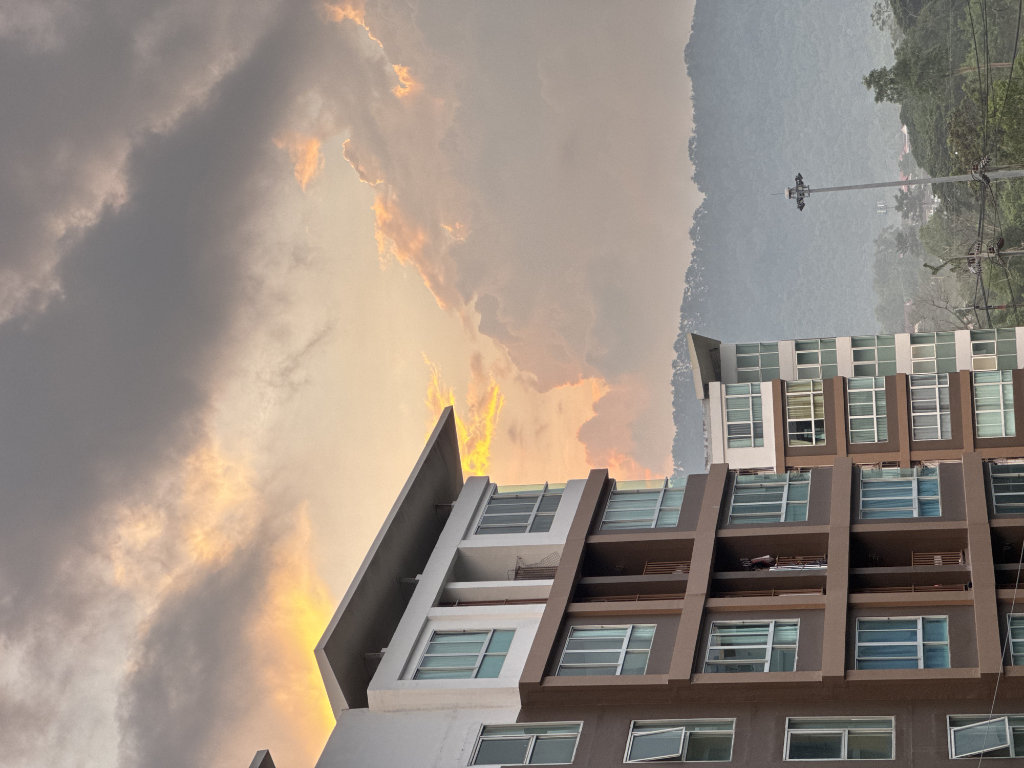The image shows a modern apartment building in Kathu, Thailand, during a dramatic sunset. The sky is filled with heavy clouds illuminated by warm orange and yellow light, creating a striking contrast with the building's architecture. The apartment building, with its multiple floors, large windows, and balconies, occupies the left side of the frame. The building's facade features a mix of brown and white sections, and some windows are open, revealing glimpses of interior blinds. In the background, a lush, tree-covered mountain range is visible, partially obscured by mist. A tall lamppost with multiple lights stands in the middle ground to the right, and power lines are present in the lower right corner. The overall scene captures a moment of natural beauty interacting with urban development.