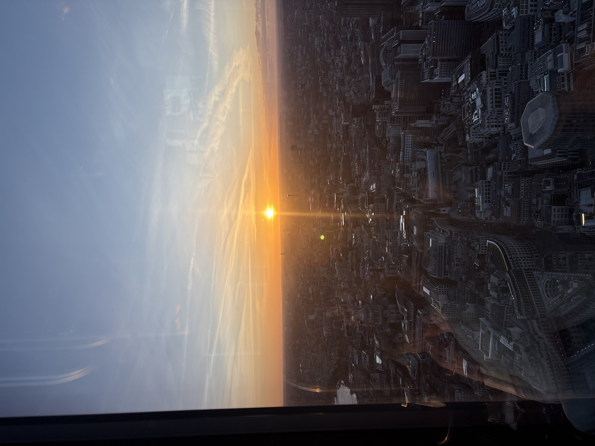 The image shows a high-angle, likely aerial view of the city of London at sunset. The sun is positioned towards the left, emitting a bright golden light that stretches across the sky and illuminates the city. A dense cluster of buildings fills the right side of the frame, with a mix of low-rise and high-rise structures, typical of London's urban landscape. The sky on the left is a pale blue, streaked with contrails. The overall impression is of a modern metropolis transitioning into twilight. The image was likely taken from a high vantage point like a skyscraper or an aircraft.