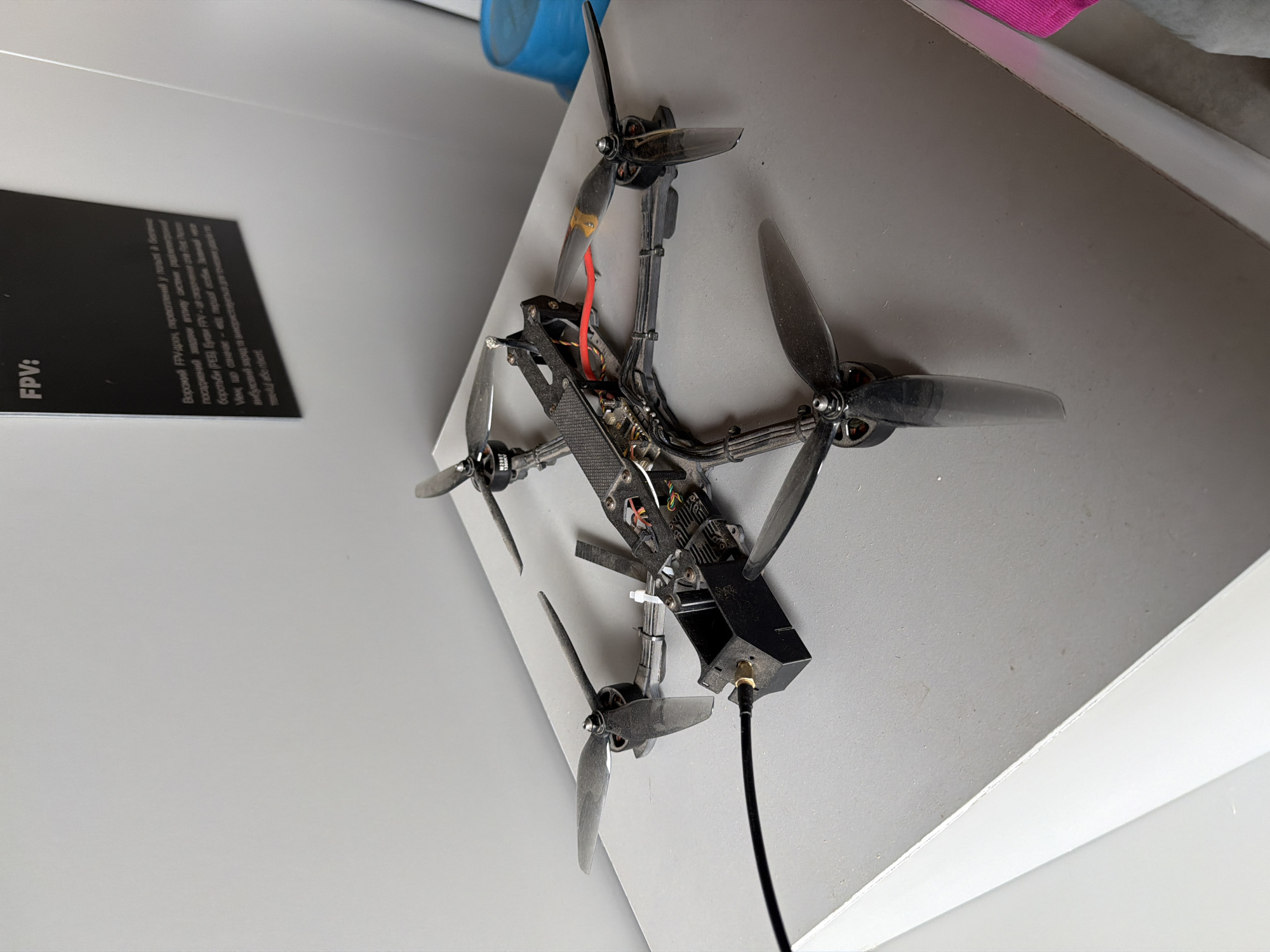 This is a forensic, matter-of-fact description of an image. The image shows a black and gray drone with four propellers, a carbon fiber frame, and various electronic components. The drone is positioned on a light gray surface, possibly a display stand, in what appears to be an indoor exhibition or museum setting. In the background, a white wall is visible, and a black plaque with text is mounted on it. The text on the plaque appears to be in Ukrainian and includes the heading "FPV:". A portion of a blue cylindrical object is visible to the right of the drone. The overall environment suggests a controlled, public display. No people are visible in the image. The location is identified as Velykyi Khodachkiv, Ukraine.