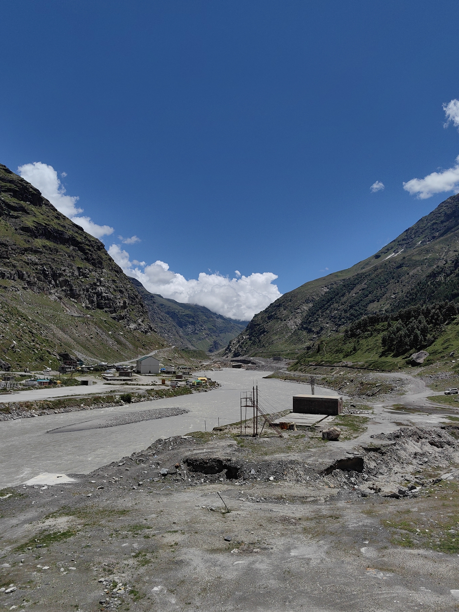 The image shows a wide, grey river flowing through a valley flanked by steep, rocky mountains with sparse vegetation. The sky is a vibrant blue with scattered white clouds. On the left side of the river, there is a small settlement with several buildings and a road. On the right side, there are more buildings, a road, and some industrial structures including a large rectangular concrete building and some scaffolding. The foreground consists of a rocky, uneven terrain with patches of dry grass. The overall impression is a remote, mountainous landscape with some signs of human habitation and activity. The location appears to be in a mountainous region of India, likely a high-altitude area given the terrain and vegetation.