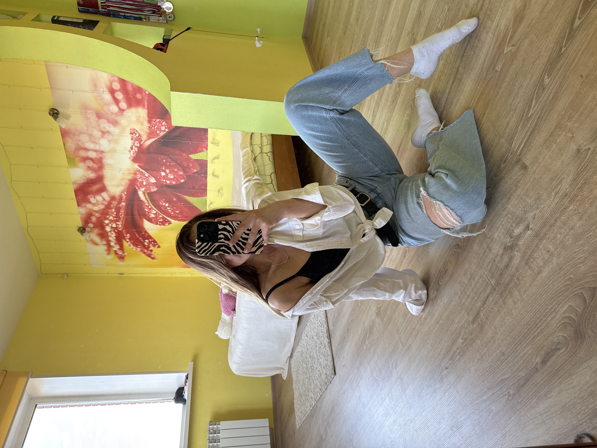 A young person, likely female, is shown taking a mirror selfie while lying on their back on a light-toned wooden floor. Their face is obscured by a black smartphone featuring a distinctive black and white zebra print case. They are dressed in a black bra, an unbuttoned white oversized shirt, light blue distressed jeans with rips at the knees and frayed hems, and white ankle socks. A delicate gold-colored necklace with a small square pendant is visible around their neck.
The setting is an indoor room, likely a bedroom or living area, characterized by bright yellow walls and a light-toned wooden floor. A prominent architectural feature is an archway decorated with vibrant wallpaper depicting a large red flower adorned with water droplets. Above this archway, a small shelf holds various items, including what appear to be books. The room also contains a white radiator situated beneath a window, which indicates natural daylight, although the view outside is overexposed. A white bed with white bedding and a white fluffy rug are partially visible.
The image captures a casual, self-posed moment in a brightly lit and decorated home interior. No specific text is discernible within the image, and the subject's emotions are not visible due to their obscured face. The provided location context is Прикамский, Russia.