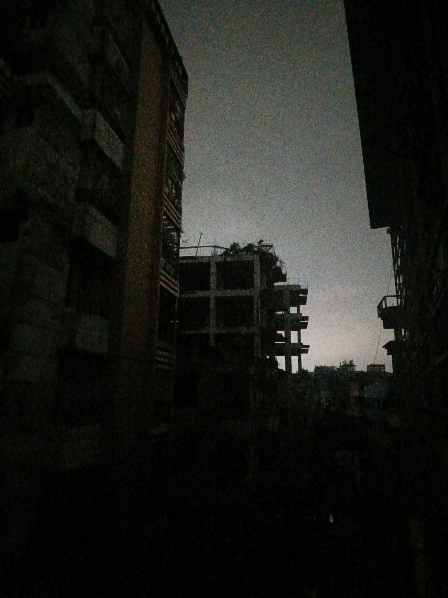 This is a nighttime, grainy photograph of an unfinished or abandoned multi-story building in Dhaka, Bangladesh. The sky is overcast and dark, contributing to the dim lighting. On the left, a portion of a dark building with visible balconies is present, partially obscuring the view of the central structure. The central structure appears to be a concrete skeleton of a building, with visible floors and structural supports, some of which are overgrown with vegetation. Another building, also appearing partially constructed or damaged, is visible on the right side of the frame, silhouetted against the sky. The foreground is largely consumed by darkness, with only faint shapes of trees or debris visible. There are no people or discernible human activity in the frame. The overall impression is one of decay and urban neglect.