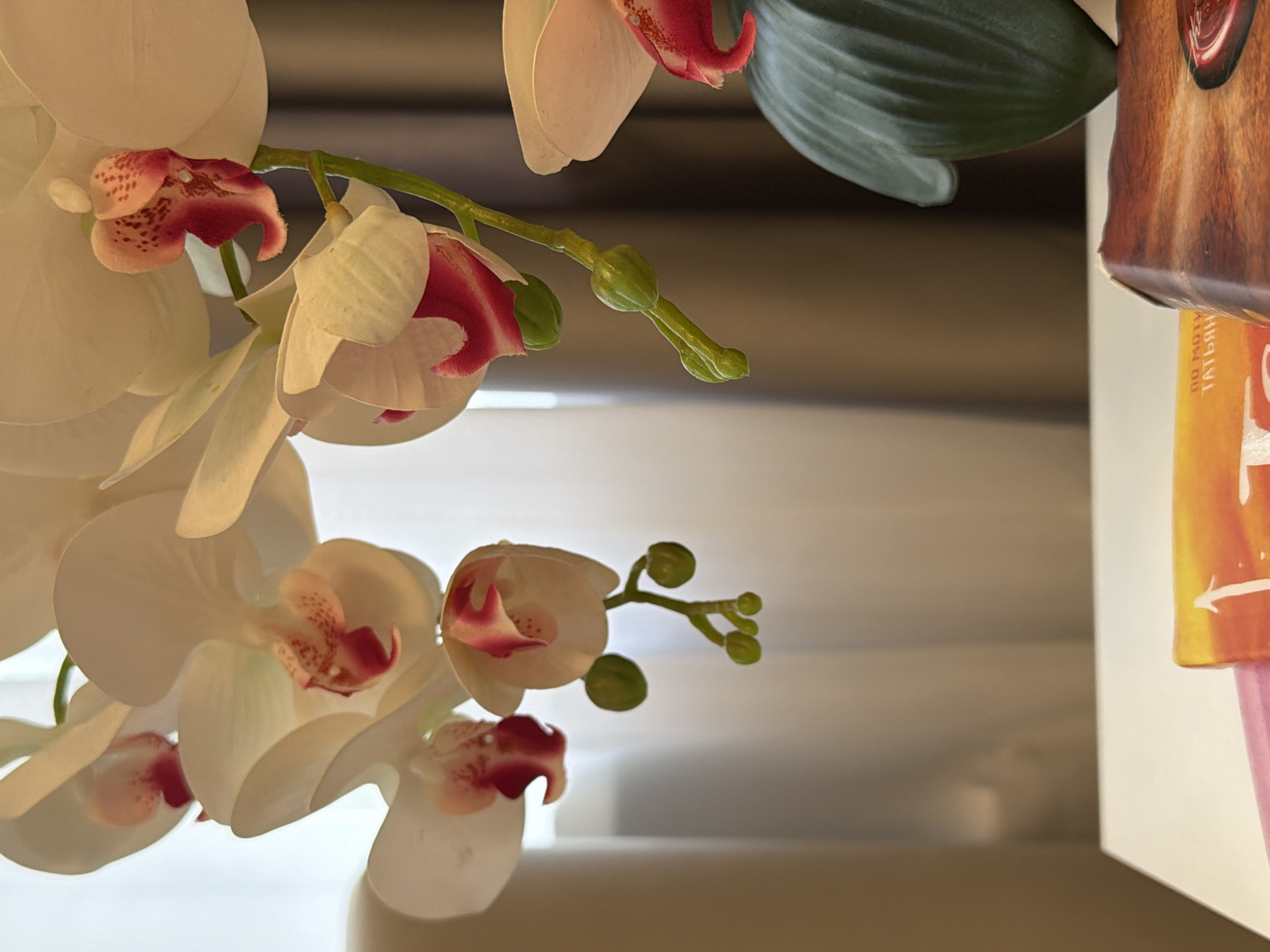 This media file features a close-up of artificial white orchids with pink centers, cascading from the top of the frame. The flowers are detailed, with delicate petals and speckled markings on the inner parts. Several buds are visible on a thin green stem, suggesting growth or a collection of blooms. The orchids are artfully arranged, with some petals overlapping and creating a sense of depth.
In the upper right corner, a portion of a decorative object is visible. This object appears to be a cylindrical item with a textured, wood-like surface and a section of colorful artwork, possibly a label or wrapper. The artwork is primarily orange and yellow, with some pink elements at the bottom. The text "ПО МОТИ" and "ТАТЬЯН" is visible in white on the orange portion of the artwork, which is oriented vertically.
The background is soft and out of focus, creating a bokeh effect. It consists of blurred horizontal lines in shades of beige and gray, possibly indicating blinds or a slatted surface. The lighting suggests an indoor setting, with soft light illuminating the orchids and casting subtle shadows. There are no people or animals visible. The overall impression is one of decorative floral arrangement in an indoor space. The mention of Kazan, Russia, provides a geographical context for the location where this media file was captured.