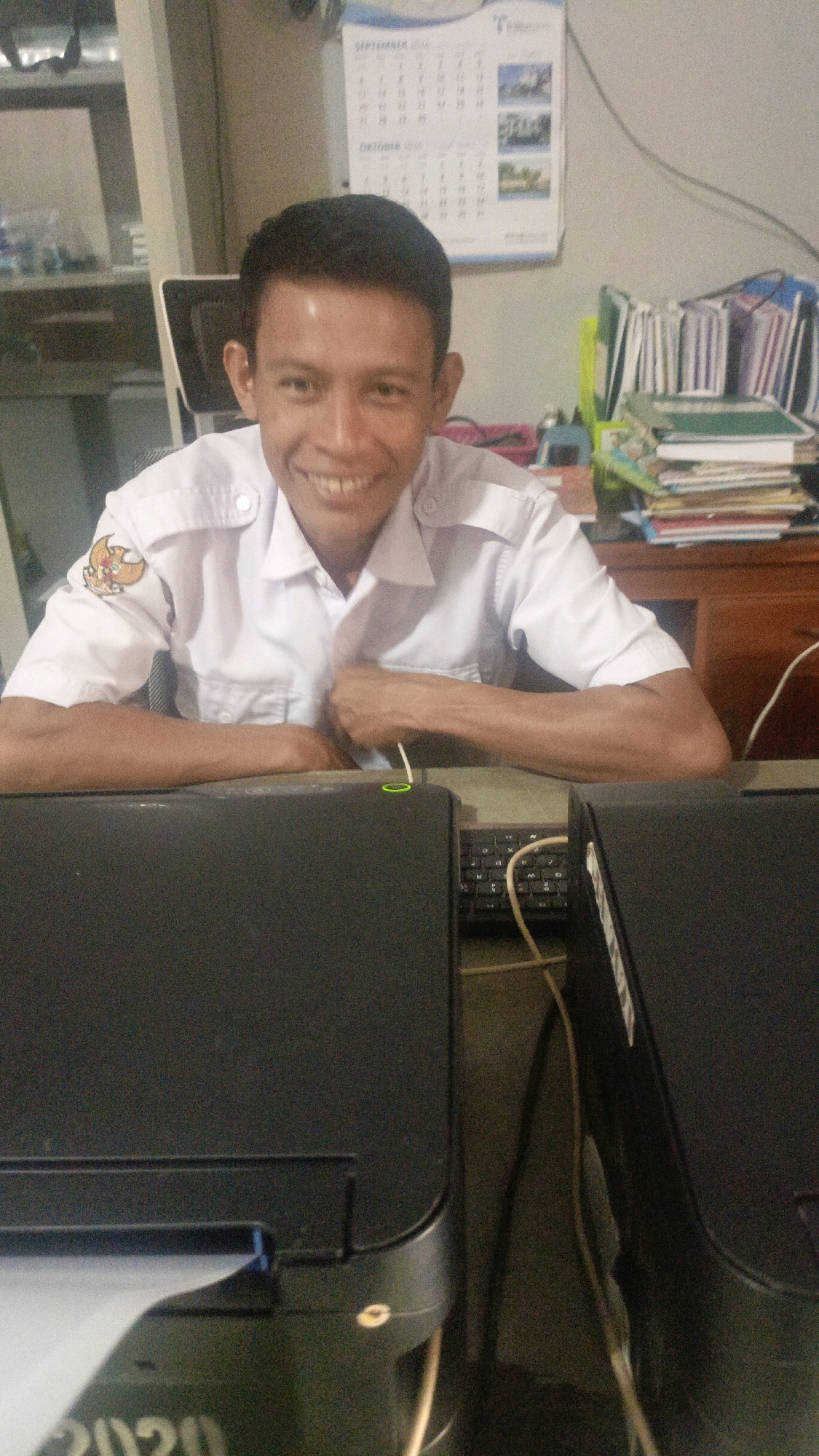 A young adult male with dark hair and a light complexion is sitting at a desk in an office environment. He is wearing a white collared shirt with a crest on the left breast. His arms are resting on the desk, and he is smiling at the camera. In the background, there is a calendar on the wall, a stack of books, and office equipment, including a printer. The scene appears to be in an indoor setting.