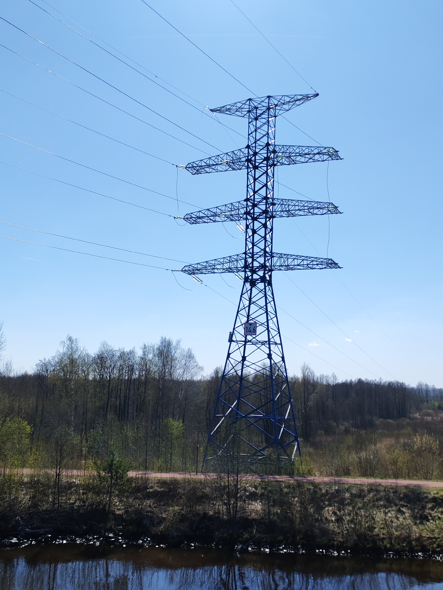A blue high-voltage transmission tower stands prominently in front of a forest and a body of water. The tower is a lattice structure with three horizontal arms holding the power lines. The sky is clear and blue. The foreground consists of a body of water reflecting the sky and surrounding vegetation. The forest in the background features a mix of bare trees and some with new spring foliage. There's a red-colored path or road visible between the water's edge and the forest. A sign with the number "14.6" is on the tower. The location in the city of Unknown, Unknown.