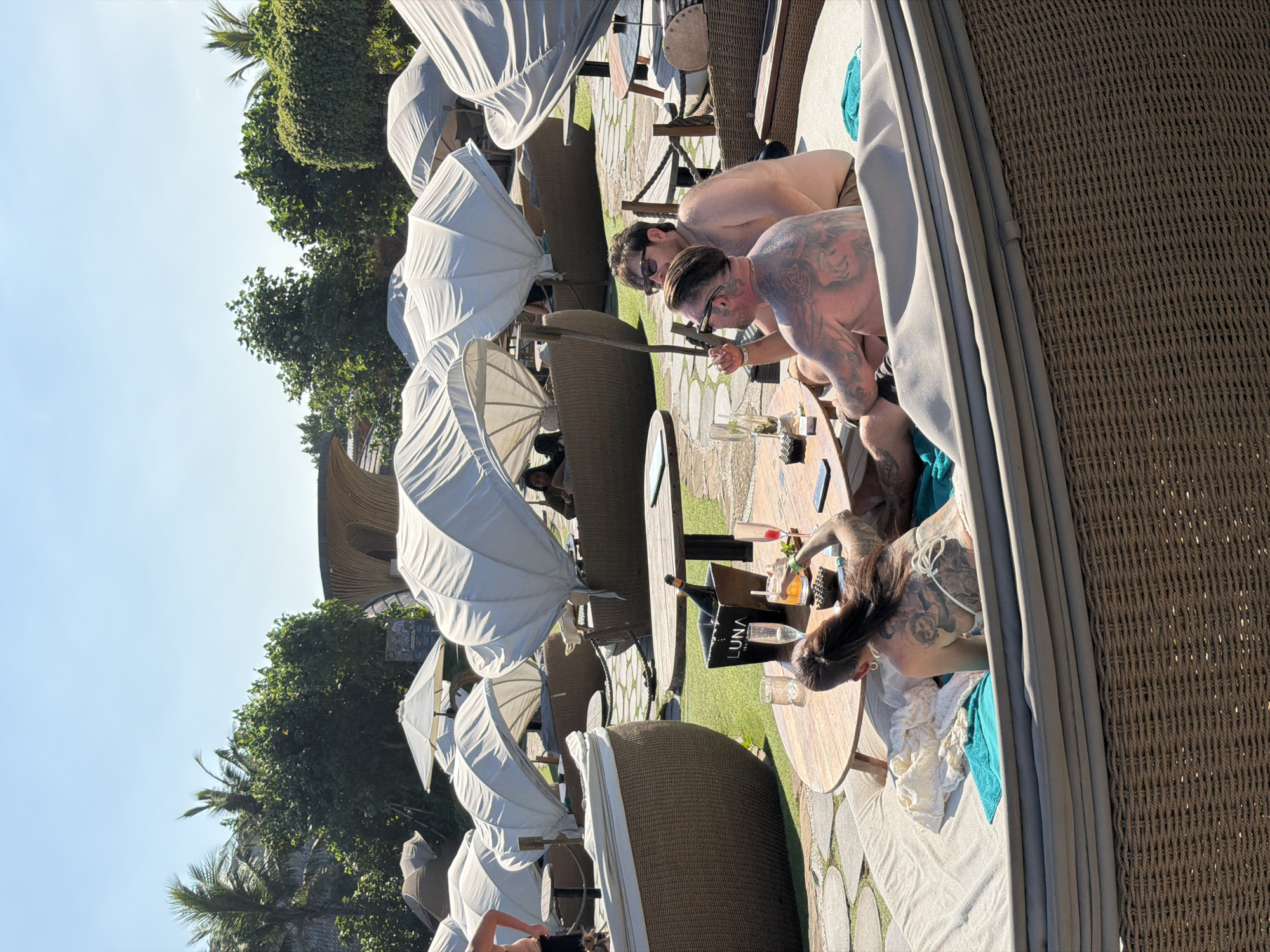The image shows a group of people relaxing at a poolside establishment in Kediri, Indonesia. The scene is set outdoors under a clear blue sky with lush green trees in the background. In the foreground, four individuals are seated around a circular wooden table. Two men, one with extensive tattoos and the other wearing sunglasses, are engaged in conversation. A woman with her back to the camera, also heavily tattooed, is seen with a drink in her hand. Another woman is partially visible in the background, seated in one of the wicker lounge chairs. On the table, there are drinks, a bottle of champagne in an ice bucket, and a phone. Several large, white canopy-covered lounge chairs are arranged around the area, suggesting a resort or a high-end beach club. The ground is paved with stone tiles, and patches of green grass are visible. The overall atmosphere is one of leisure and relaxation.