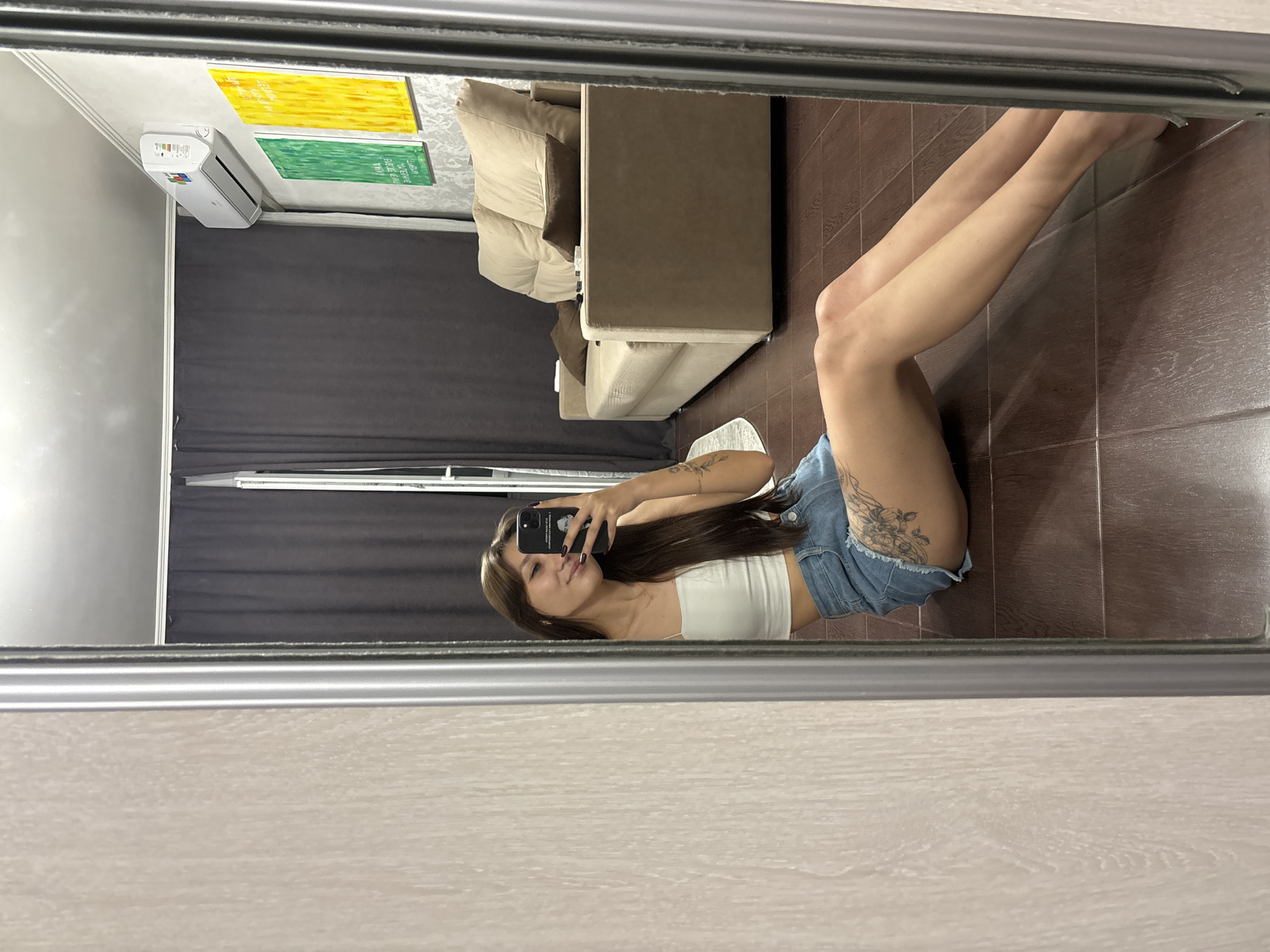 A young woman with long brown hair is taking a mirror selfie in an indoor setting. She is wearing a white crop top and denim shorts, and is sitting on a dark wood-tiled floor. She is holding a black smartphone in her right hand. In the background, a beige sofa, a wall with two abstract paintings (one green, one yellow), and a white air conditioning unit are visible. The scene suggests a domestic environment, possibly an apartment. The user has provided no information about the location of this image within the city of Novokuznetsk, Russia.