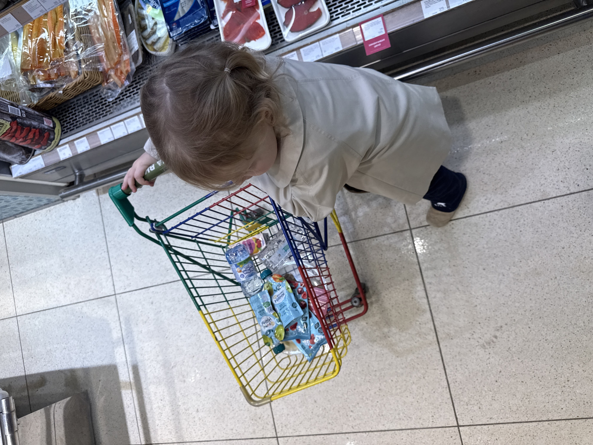 An overhead photograph captures a young child pushing a multi-colored toy shopping cart within a supermarket aisle. The child, with light brown hair pulled into a small ponytail, wears a light-colored coat and dark socks with light-colored slippers. Their gaze is directed downwards, possibly at the contents of their cart.
The toy shopping cart is filled with several items, including small bottles of "Smalis" water and fruit puree pouches, some visibly branded "NANA DUCK" and "Чудо Чадо," with "100%" printed on them.
The setting is a well-lit supermarket, identifiable by the tiled, light-colored floor and refrigerated display cases lining the aisle. These cases hold various packaged food items, such as smoked fish and red fish fillets. Visible price tags on the shelves display prices in rubles, like "319р" and "205р," along with Cyrillic text that includes "ЗАМОРОЖЕННЫЙ" (Frozen) on one label, consistent with the location of Moscow, Russia. The image portrays a common scene of a child imitating adult shopping behavior.