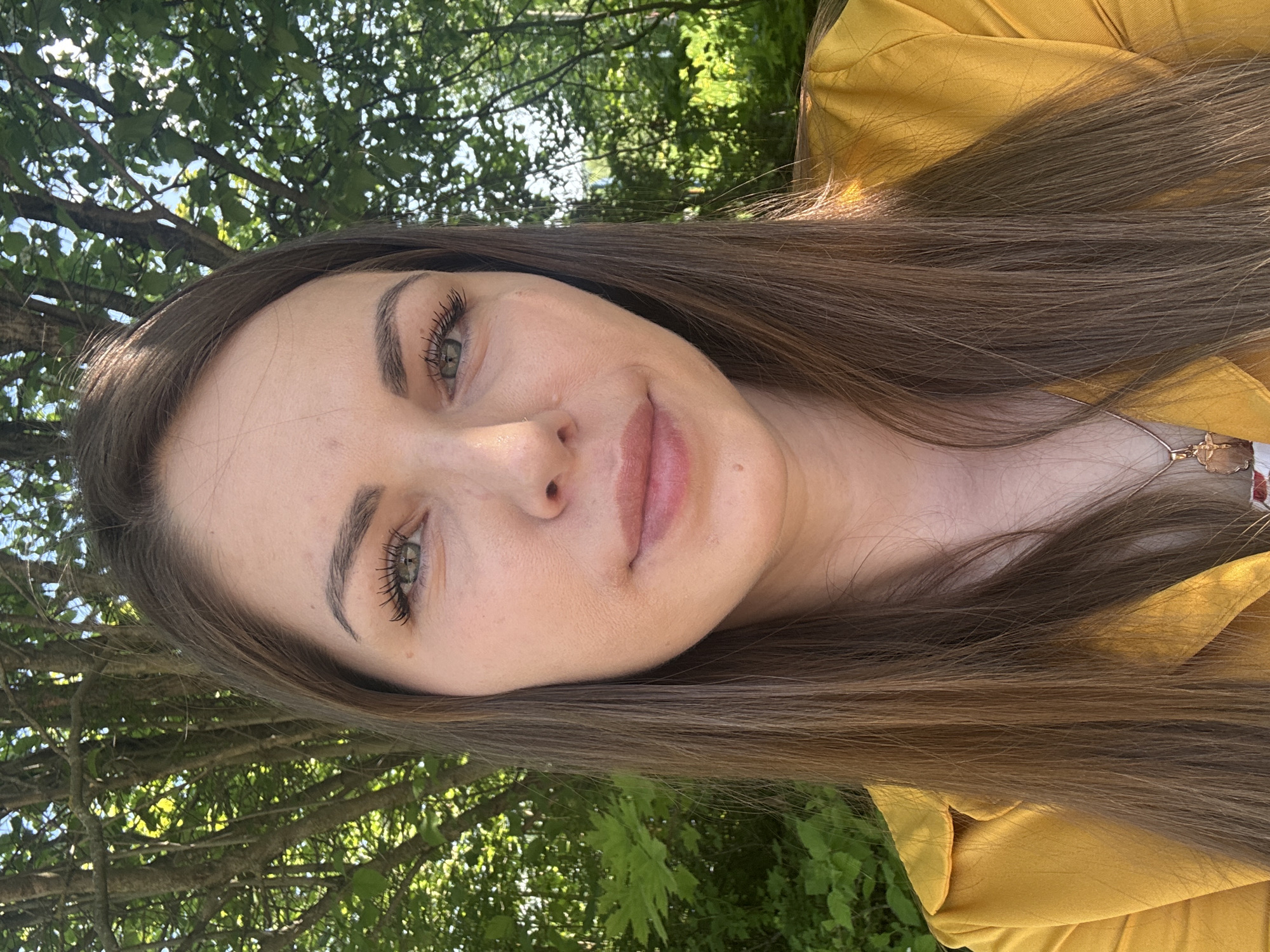 A woman with long, straight brown hair is wearing a mustard-yellow jacket and taking a selfie outdoors. The background consists of green foliage and tree branches against a bright sky. There is no evidence of this image being related to Shevlyakovo, Russia.