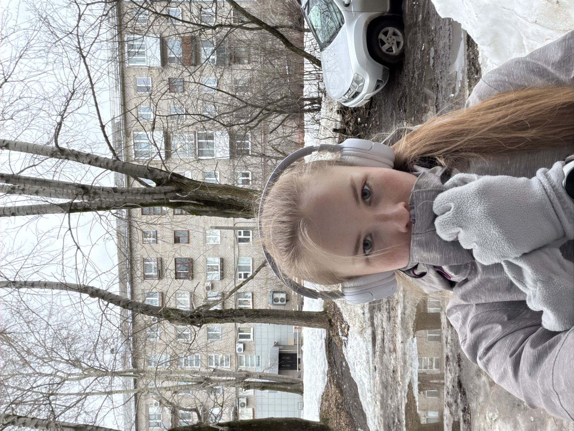 A young person with headphones stands outdoors in winter. Snow remains on the ground. In the background, there is a multi-story apartment building constructed of grey brick. A bare tree and a white car are also visible in the background. The apartment building is in Berezniki, Russia.