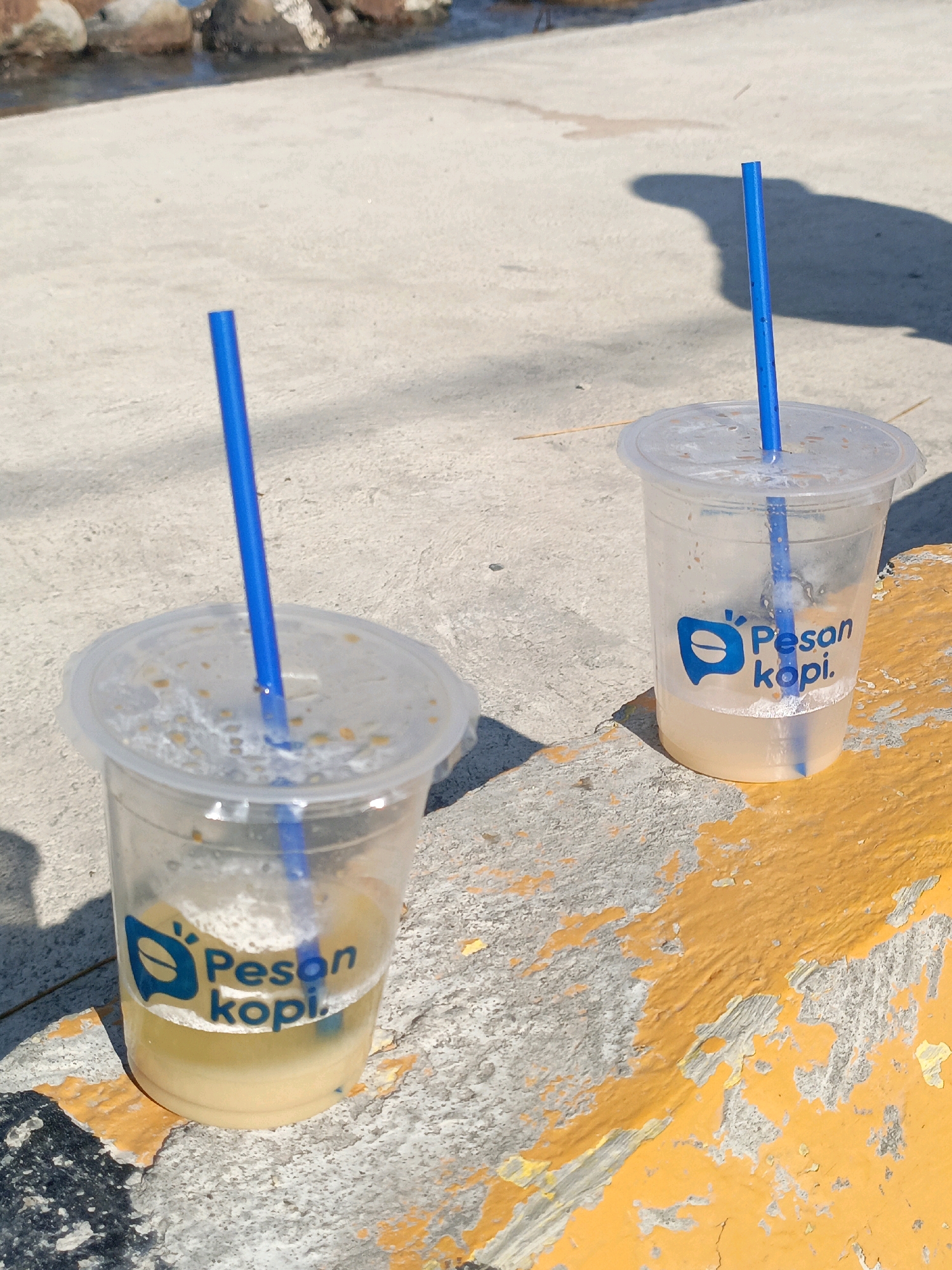 Two clear plastic cups with blue straws and the logo "Pesan kopi." are placed on a concrete surface with some yellow paint peeling off. The cups contain a light-colored liquid, and one cup has a small amount of liquid remaining. The background shows a body of water with rocks. The scene is likely outdoors in Bulukumba, Indonesia, given the context of "Pesan kopi.", a local coffee shop. The lighting suggests it is daytime. There are no people visible in the image.