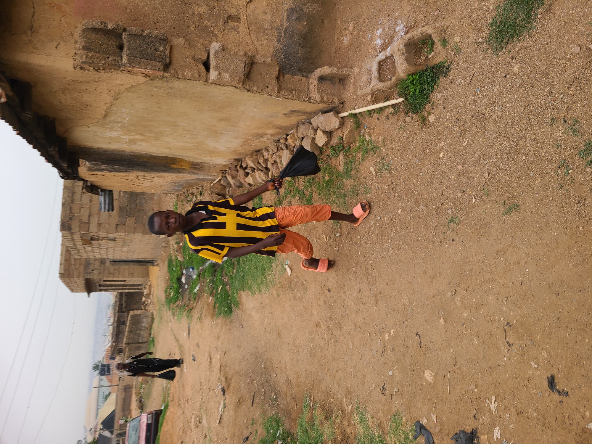 The scene is an outdoor environment located in a neighborhood in Jos, Nigeria. The ground is dirt and gravel with scattered patches of grass. To the left is a weathered building constructed from blocks of clay or concrete, with visible deterioration and dark streaks on its exterior.
A young person is standing on the pathway. The person is wearing a black and yellow striped t-shirt, bright orange shorts, and orange sandals. The person is holding a black plastic bag. In the background, another person can be seen walking farther down the dirt path. A car and other buildings are also visible.