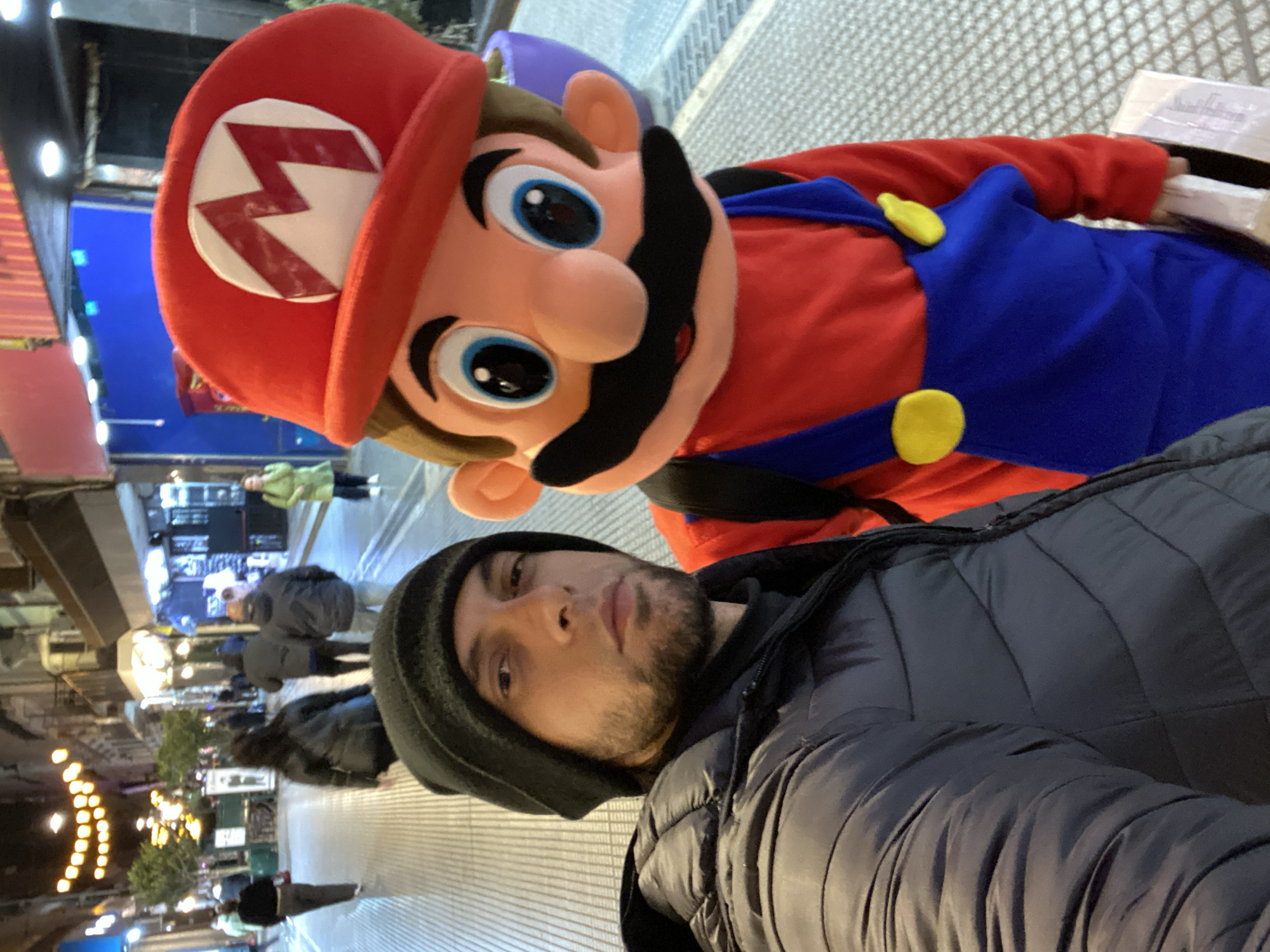 A man in a black beanie and puffy black jacket takes a selfie with a person dressed as Mario from the Super Mario Bros. video game franchise. They are positioned on a pedestrian street with lit storefronts and streetlights in the background, with other people walking in the distance. The street is lined with light-colored pavers. The image was taken at night in the city of Buenos Aires, Argentina.