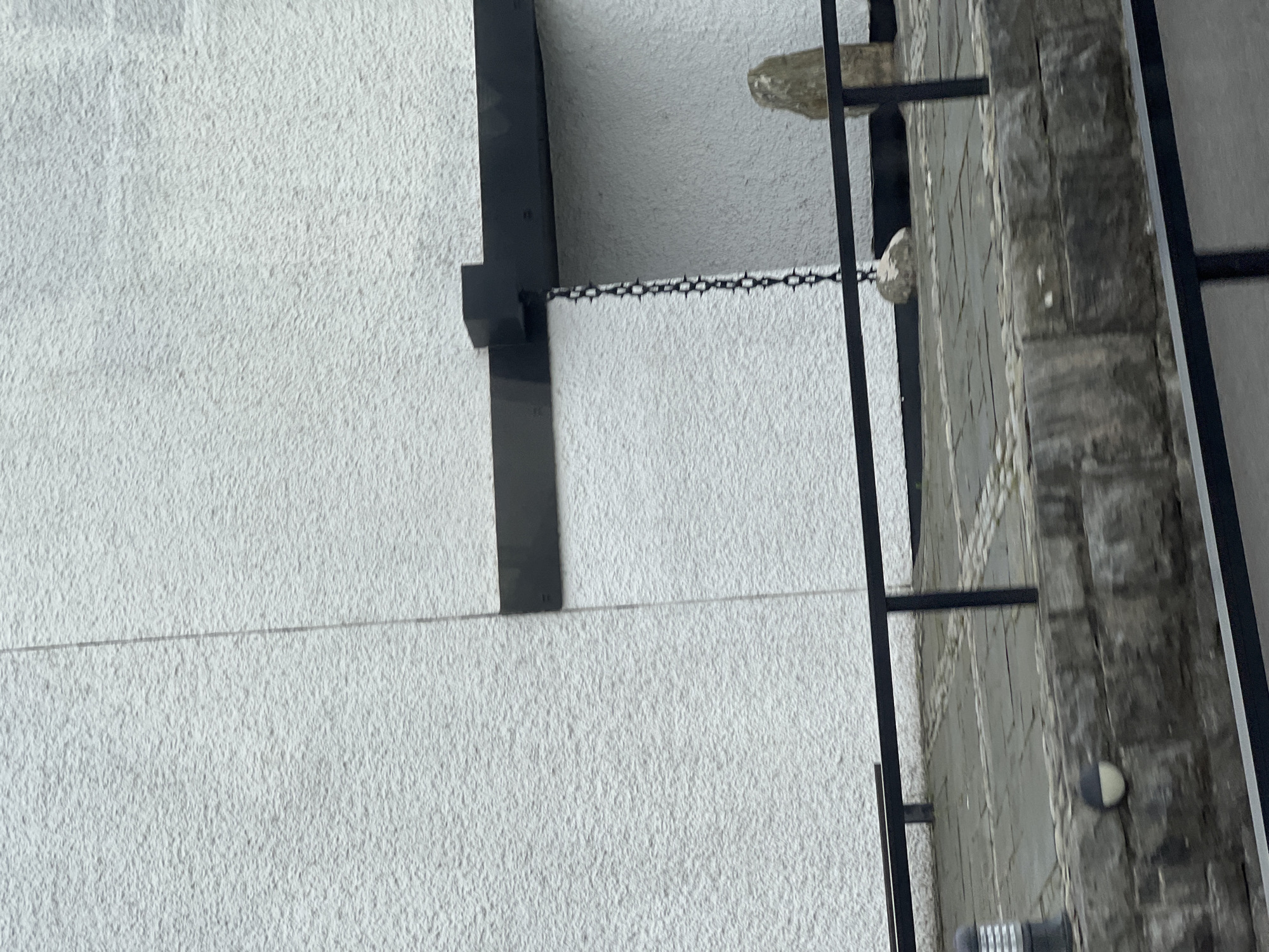 The image shows the exterior of a building in Creeslough, Ireland. A textured white wall dominates the background, with a dark horizontal beam extending from the upper right of the frame. Hanging from this beam by a chain is a rock. To the right of the hanging rock, a larger, rough-hewn stone stands upright. In the foreground, a black metal railing runs horizontally across the scene, separating a paved walkway from a stone wall below. The walkway is made of grey flagstones and cobblestones. A cylindrical light fixture with silver and grey elements is visible on the left side of the walkway.