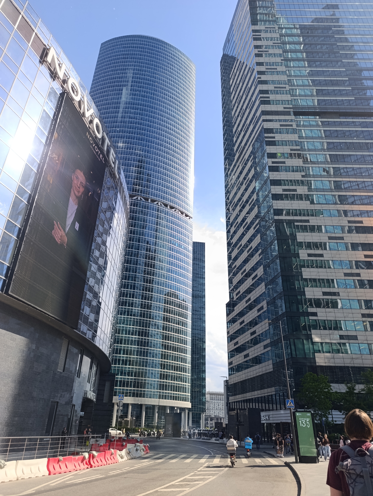 The image captures a vibrant urban scene in Moscow, Russia, dominated by several towering, modern glass skyscrapers under a bright, clear blue sky with a few wispy clouds. The time of day appears to be midday or early afternoon, with strong sunlight reflecting off the glass facades.
In the foreground and to the left, a curved glass building, identified as "NOVOTEL" by its large signage, features a prominent LED screen displaying a portrait of a man. The building's reflective surface shows a clear blue sky and sun glare. To the right of the Novotel, a massive, cylindrical skyscraper, entirely clad in blue-tinted glass panels, rises majestically into the sky. Further right, another angular skyscraper with a more complex pattern of glass and darker gray panels also reflects the surroundings.
At street level, traffic barriers line the road, which features pedestrian crossings. Numerous people are visible, engaged in various activities. A person on a small scooter or moped and another on a bicycle are seen on the road. Several pedestrians are walking on the pavement, including a person in the foreground with a backpack, their back to the camera.
Notable details include a bus stop or information sign displaying the text "БУЛГАКОВ 135" and "ЗБАНК", indicating a specific street or location reference. A pedestrian crossing sign is also visible. The overall scene conveys a bustling, modern city environment, characteristic of a business or commercial district.