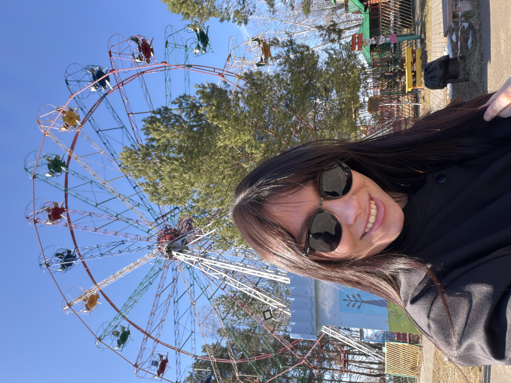 The image captures a bright, outdoor selfie of a woman with long, dark hair, wearing sunglasses and smiling cheerfully at the camera. She is positioned in the foreground, slightly angled, with a vibrant amusement park setting behind her.
The dominant feature in the background is a large Ferris wheel with a white metal structure. Its numerous gondolas are brightly colored in shades of red, yellow, green, and blue, some appearing to be empty. The sky above is a clear, cloudless blue, indicating a sunny day. Scattered around the Ferris wheel and in the mid-ground are several lush green trees, likely conifers, suggesting the location is a park or recreational area.
To the right of the woman, further into the background, elements of an amusement park are visible. These include a small building with a green roof, possibly a kiosk or ticket booth, fronted by a fence and yellow park benches. A black trash bag is visible in a bin near the benches. A sign attached to the green-roofed building displays Cyrillic text: "СПАСИБО ЗА ЧИСТОТУ!" (Spasibo za chistotu!), which translates to "Thank you for cleanliness!" The sign also features two small, light-colored figures, possibly cartoon bears.
The overall impression is one of a pleasant, sunny day spent at an amusement park. The woman's smile conveys happiness and relaxation. Given the provided context, the scene is located in Chaikovsky, Russia.