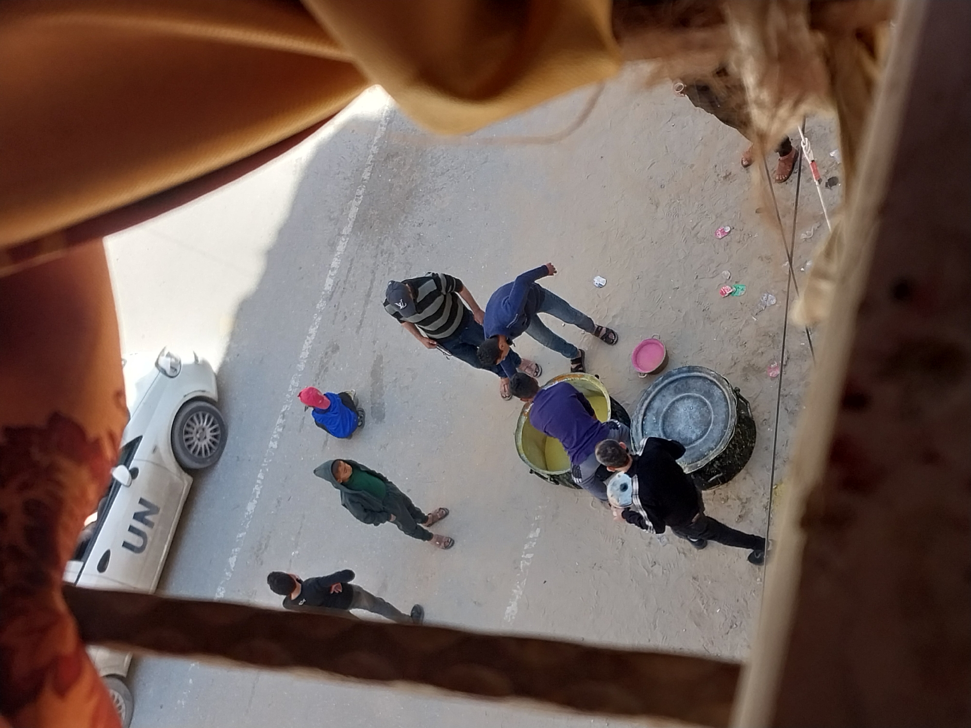 The image, taken from an elevated perspective, shows a group of people in what appears to be a city street. A white vehicle with "UN" markings is parked on the left side of the image. Several individuals are gathered around large containers, possibly drums or barrels, some of which seem to contain a yellow liquid. One person is actively interacting with the containers. Other people are standing nearby, and their clothing suggests they are civilians. The street surface is plain, and some debris or objects are scattered near the containers. The overall setting suggests a community activity or interaction involving the United Nations vehicle.