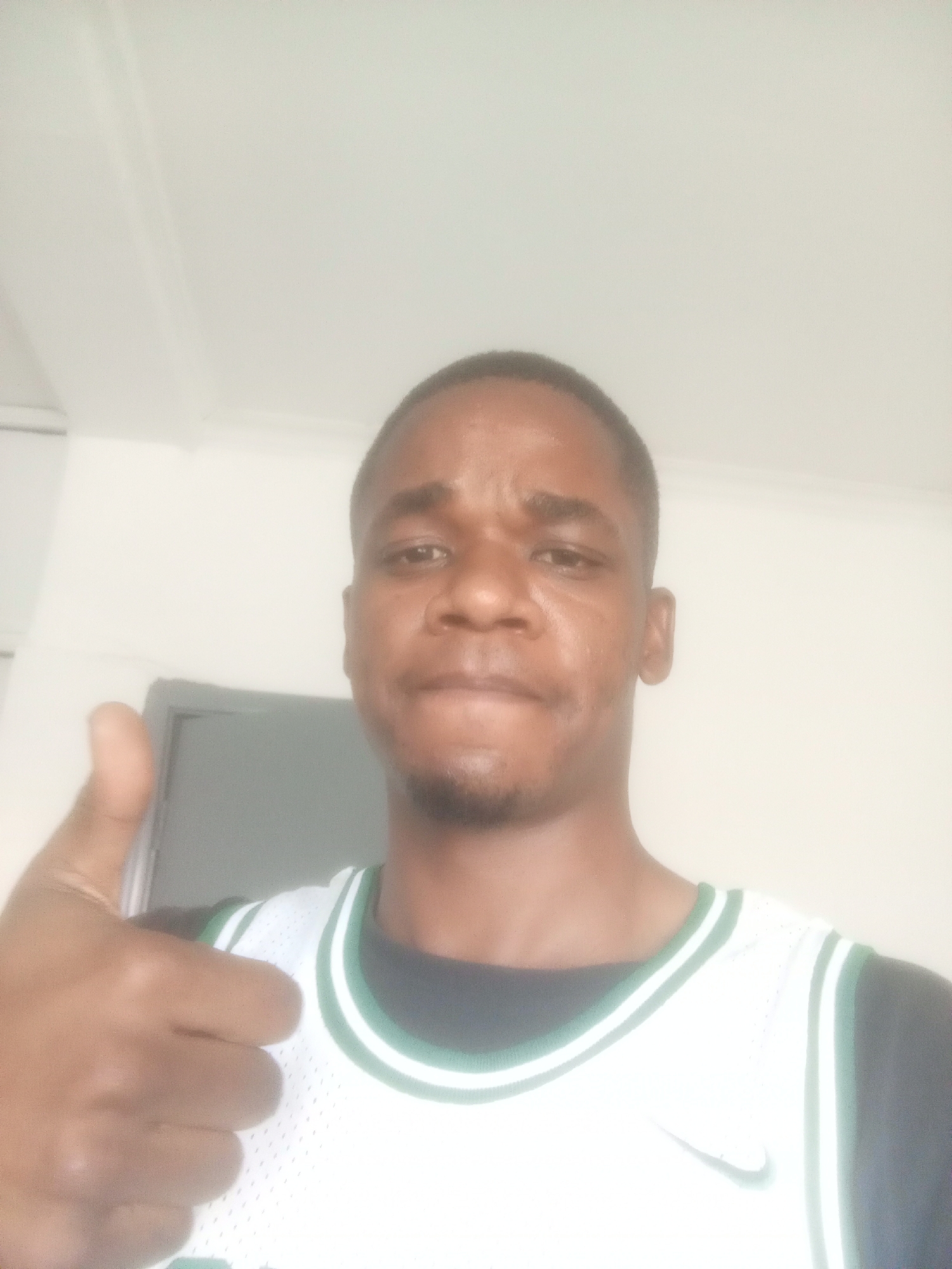 A close-up shot of a man, likely in his 20s or 30s, giving a thumbs-up. He has short, dark hair and a slight beard. He is wearing a white basketball jersey with green trim over a black undershirt. The background is a plain white wall. A gray object resembling a television is partially visible behind him. The image was likely taken indoors. There is insufficient information to determine the specific location in Abuja, Nigeria.