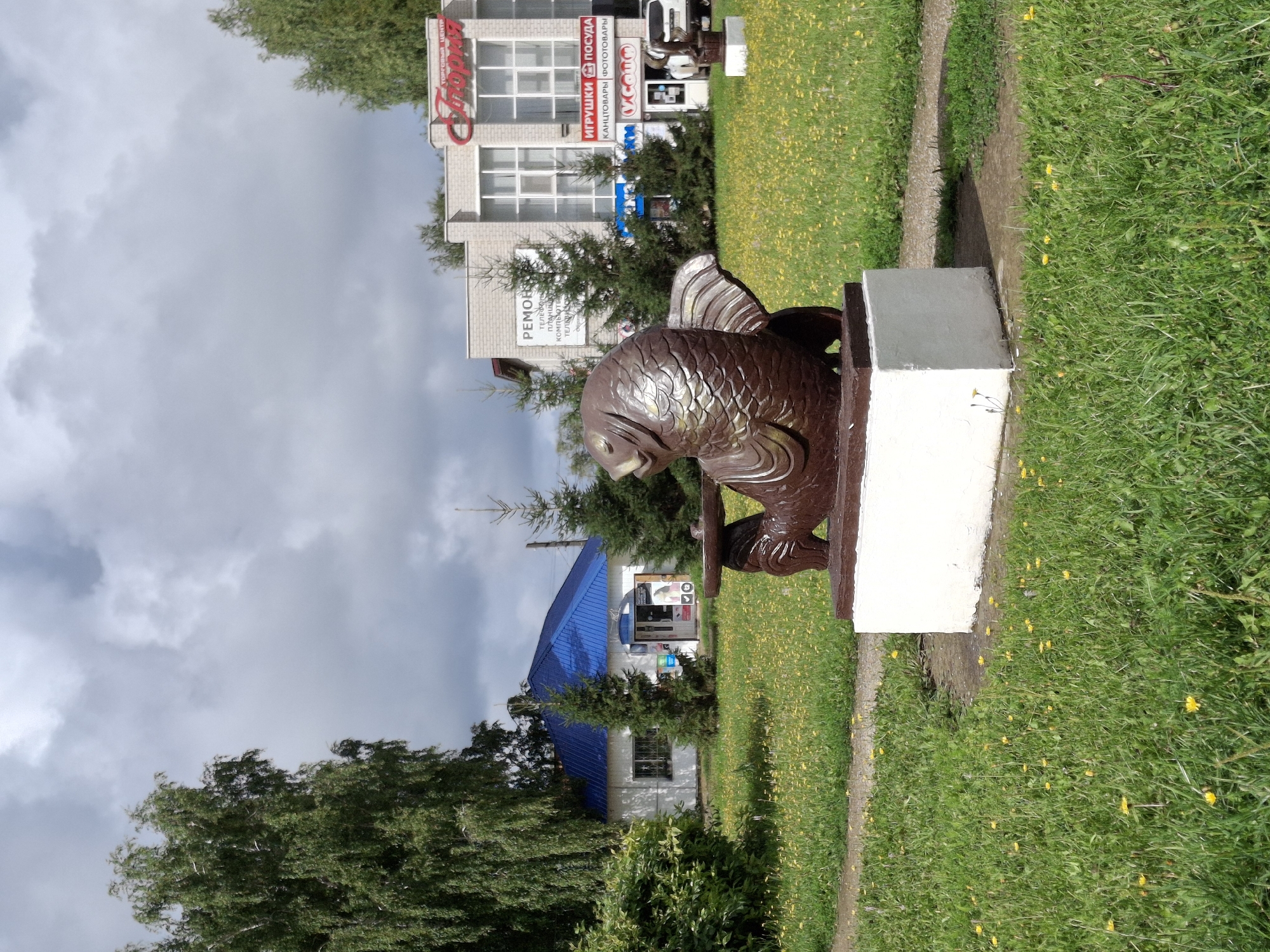 In the city of Elovo, Russia, a brown, fish-shaped sculpture sits on a white pedestal in a grassy area dotted with yellow flowers. The sculpture appears to be made of concrete or a similar material, with a textured surface mimicking scales. It is positioned on the left side of the frame, facing to the right. Behind the sculpture, a white building with a bright blue roof is visible, featuring several windows and an open doorway. To the right of this building, a larger, light-colored brick building displays several signs. One prominent sign in red letters reads "Торговый Центр" (Shopping Center) followed by "Глория" (Gloria). Another sign indicates "ИГРУШКИ ПОСУДА" (Toys, Tableware), and below that "КАНЦТОВАРЫ ФОТОТОВАРЫ" (Stationery, Photo Goods). A white SUV is parked in front of this building. The sky above is cloudy with patches of blue. Tall green trees frame the scene on the left and in the background. The overall impression is of a public space within a small town or city.