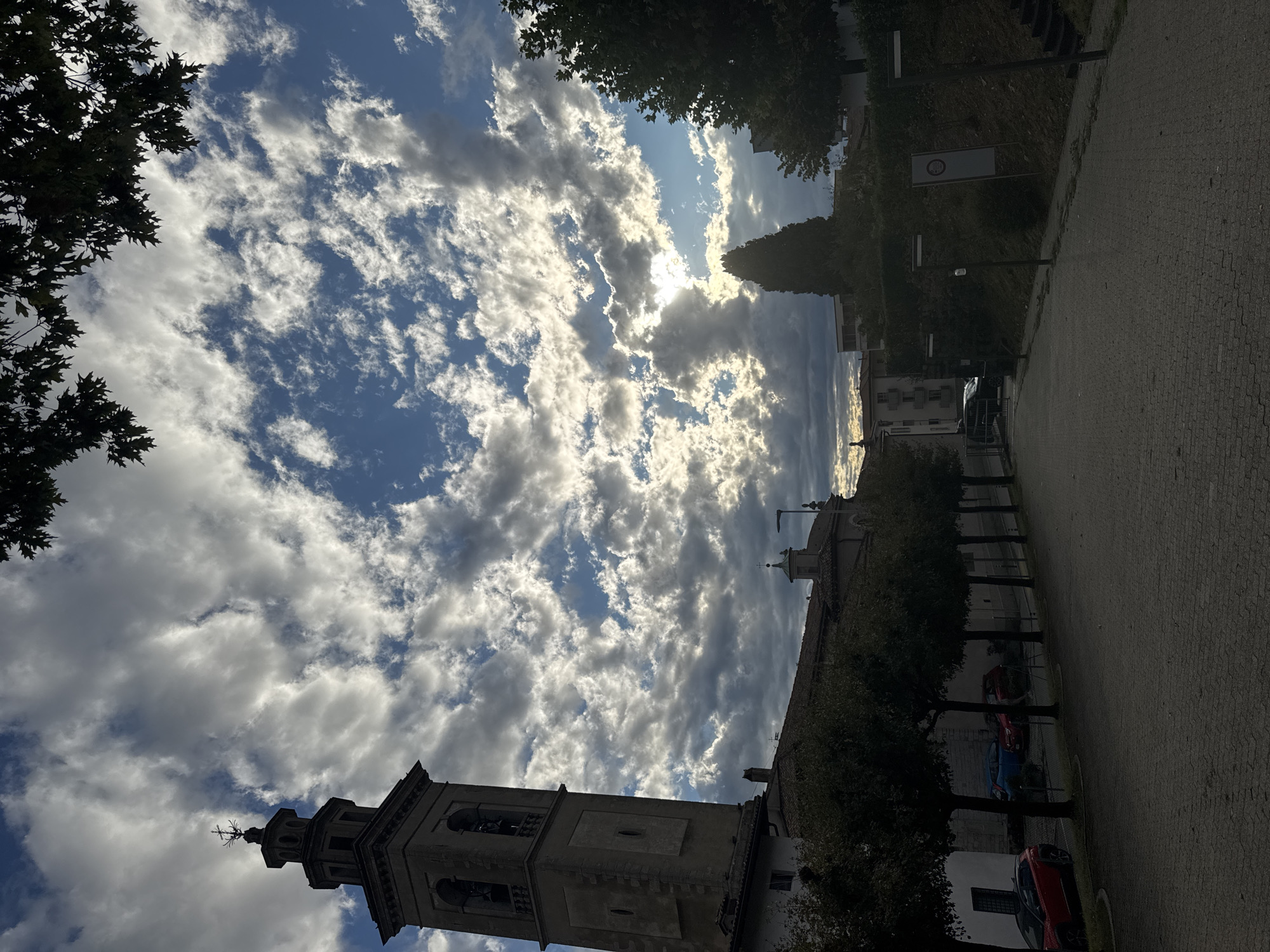 A street view in Balerna, Switzerland, shows a bell tower to the left, a paved walkway in the foreground, and trees lining the right side of the walkway. The sky is filled with dramatic clouds, with sunlight breaking through in the distance. Several parked cars are visible behind a fence to the left. The scene appears to be set in the late afternoon or early evening, with shadows lengthening.