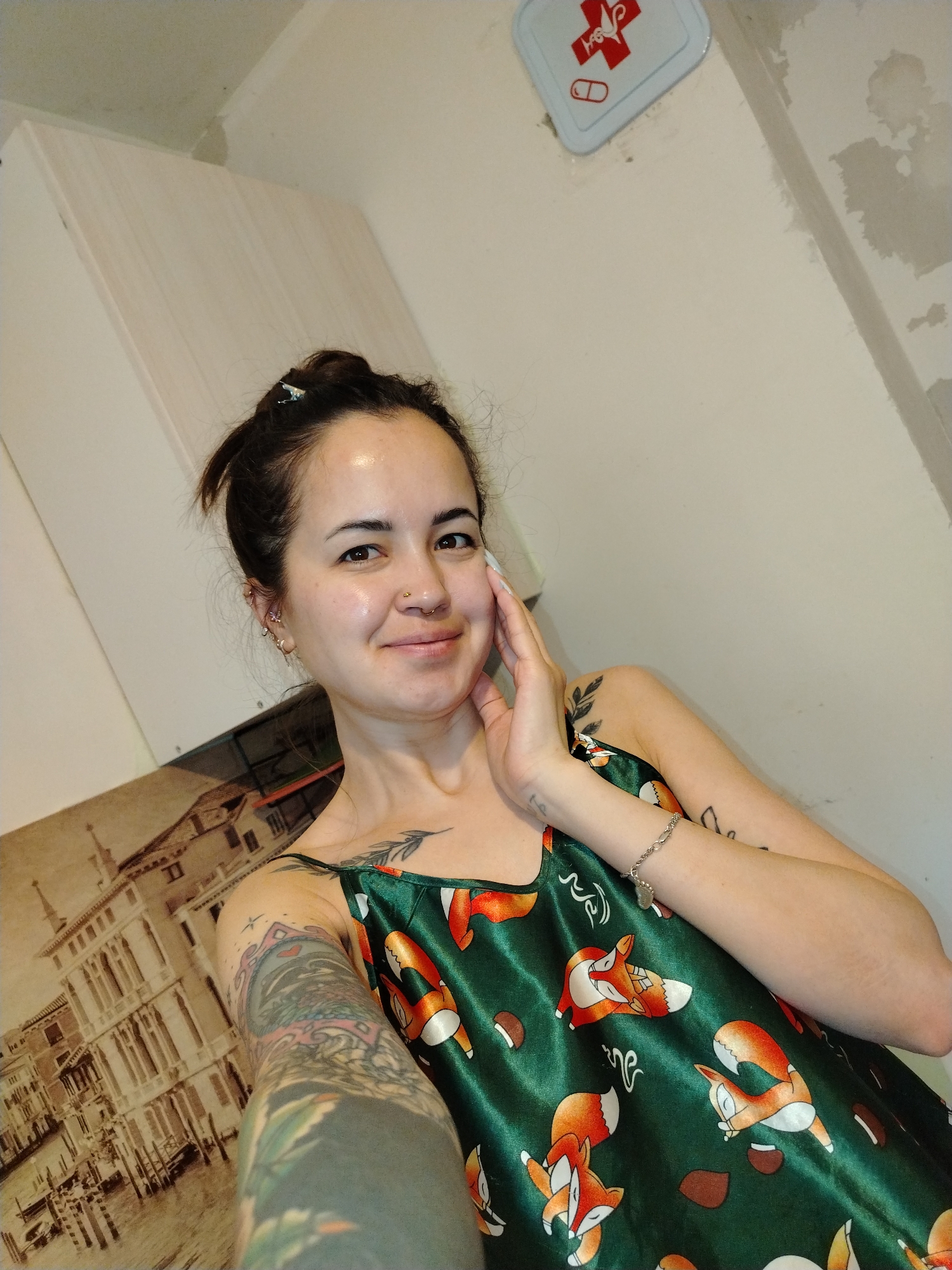 A young woman, likely in her 20s or 30s, takes a close-up selfie in what appears to be a casual indoor setting in Noviy, Russia. She has dark hair pulled back into a bun with a small light-colored clip, dark eyes, and a gentle, relaxed smile, looking directly at the camera. Her complexion is natural, and she has several visible piercings, including a septum ring, a nostril piercing, and multiple ear piercings (helix, tragus, and lobe).
She is wearing a dark green, shiny camisole-style top, which is patterned with cartoonish orange and white foxes in various poses, along with brown acorns or chestnuts. Her left arm, extended towards the camera, is adorned with an extensive, detailed sleeve tattoo. A smaller, decorative tattoo of leaves or branches is visible on her right collarbone/shoulder area. She also wears a simple silver chain bracelet on her right wrist, while her right hand gently rests against her cheek.
The background shows a mix of functional and decorative elements. To her upper right, a small white first aid kit is mounted on a light-colored wall, featuring a red cross symbol and a red pill icon. The walls appear to be painted in a light, neutral tone, with some areas showing rougher texture or patches, suggesting an unfinished or utilitarian space. To her upper left, a light wooden cabinet or panel is visible. On the lower left side, behind her arm, a large, sepia-toned picture or tapestry depicting buildings along a canal, possibly Venice, is partially visible. The lighting is bright and even, consistent with an indoor environment.