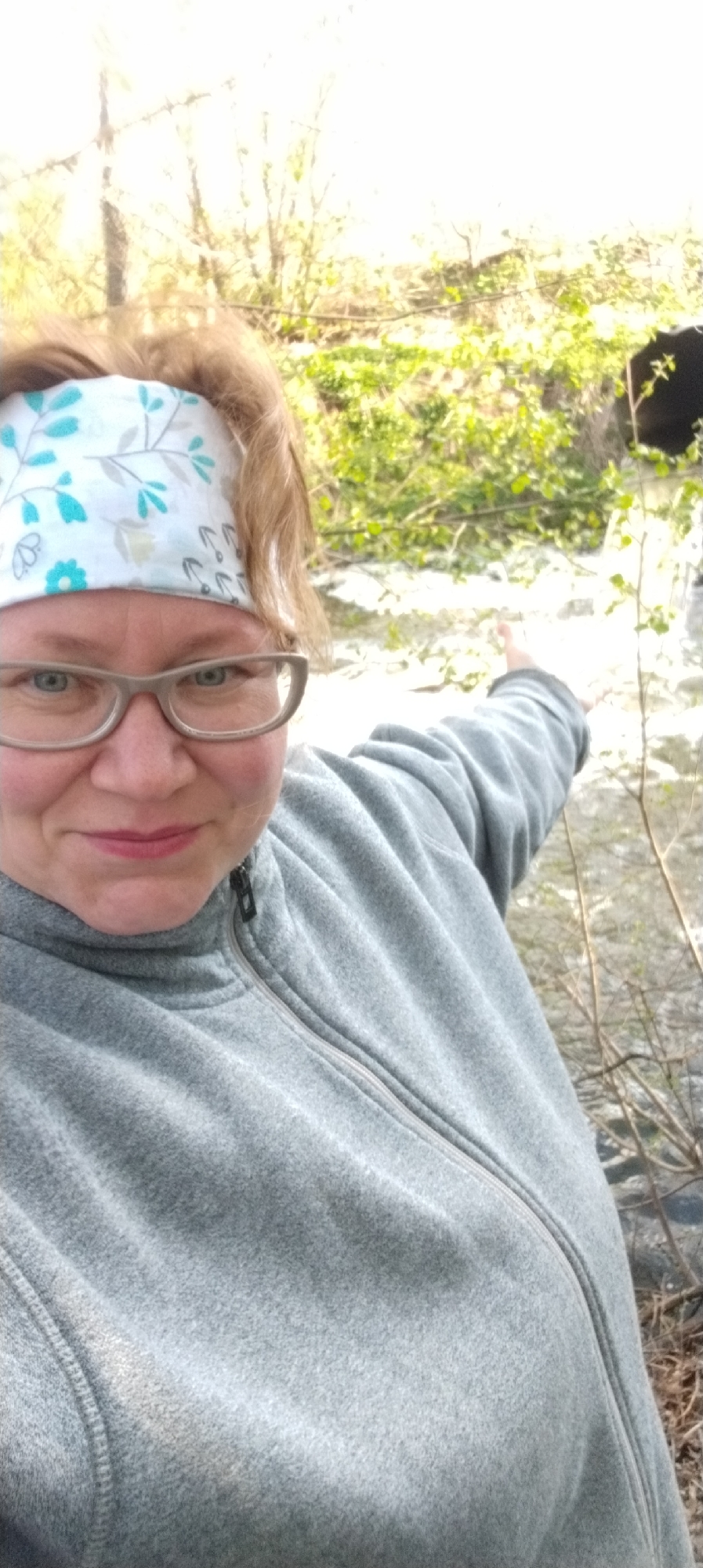 A selfie shows a middle-aged woman wearing glasses and a floral headband, pointing towards a dark opening surrounded by greenery in Зипуново, Russia. She is wearing a gray fleece jacket. The background includes foliage and a light-colored surface, possibly a creek or rocks. The lighting is bright, suggesting daylight. The focus is primarily on the woman and the immediate area she is pointing towards.