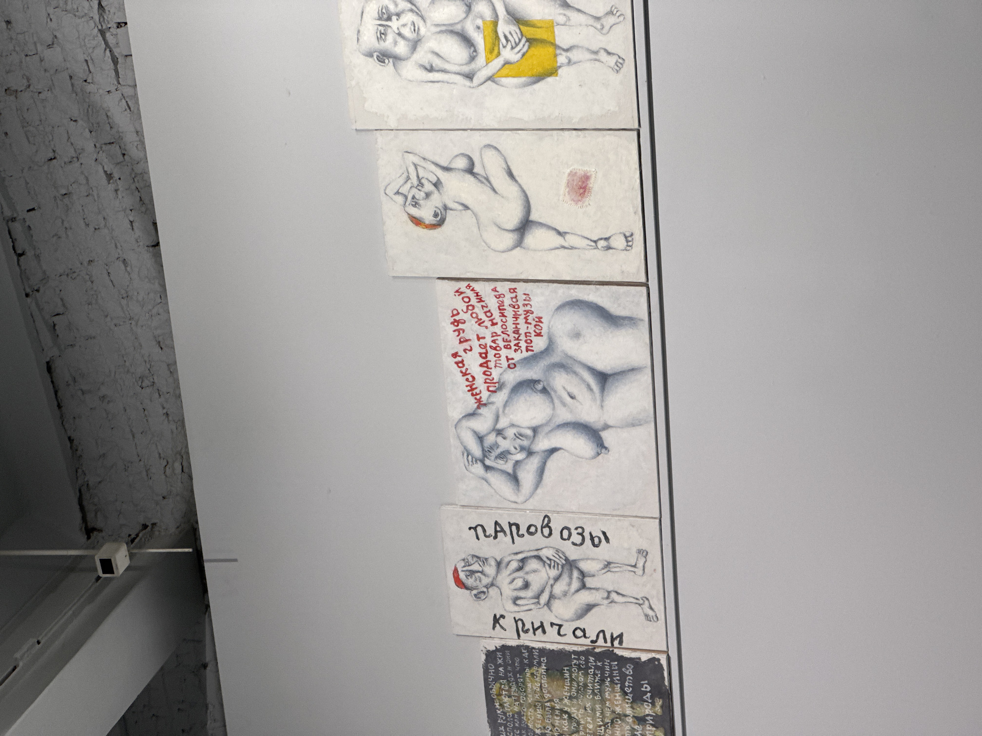 The image shows a vertical arrangement of several small artworks, likely paintings or drawings, on a white wall, possibly in a gallery or exhibition space in Moscow, Russia. The artworks depict nude figures in various poses and also have some Russian text accompanying them. The artwork near the bottom is captioned with the word "паровозы," which translates to "locomotives," and another word, "кричали", which translates to "shouted." The artwork immediately above features Russian text describing the female body. The top three paintings feature nude or partially clothed figures. A brick wall is visible to the left. A security camera can be seen on the ceiling on the left.