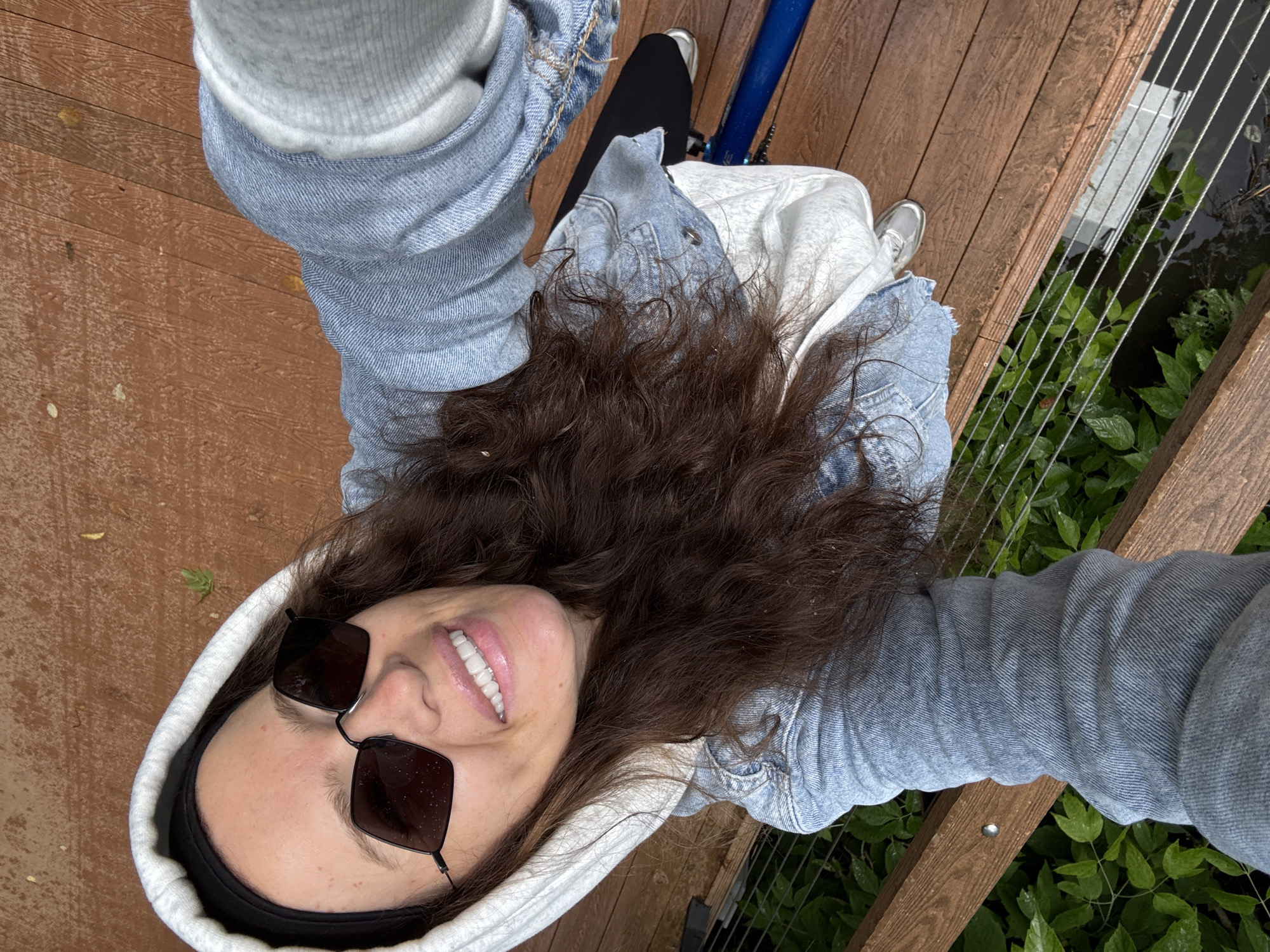 In a high-angle, selfie-style shot, a woman with long, dark brown hair is pictured outdoors. She is wearing a light blue denim jacket over a white hooded sweatshirt, a black headband, and dark, angular-shaped sunglasses. The woman is smiling broadly, revealing white teeth. She appears to be standing on a wooden deck, with green foliage visible below and to the left. The background consists of weathered wooden planks and a railing with thin metal wires. There is a dark, possibly black object, resembling a skateboard or similar item, visible beneath her. A bright blue cylindrical object is also partially visible. The scene is overcast, and there are visible water droplets on the sunglasses and foliage, suggesting recent rain. The woman is positioned in the center of the frame, with her head tilted upwards towards the camera. The overall impression is casual and relaxed. There is no information to indicate this scene is located in Moscow, Russia.