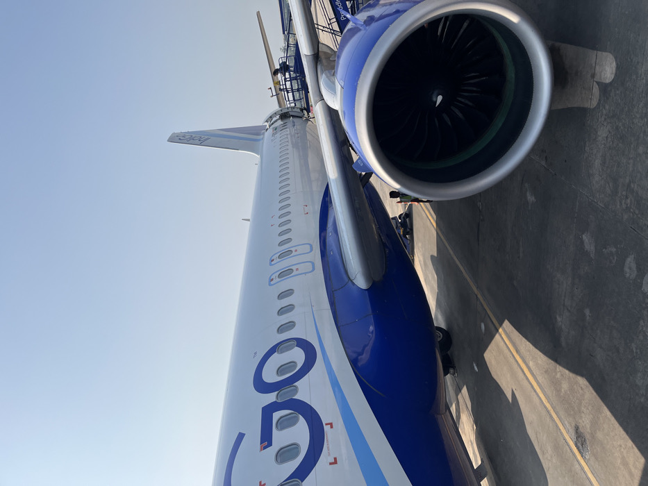 The image captures a ground-level, slightly upward-looking view of a white and blue passenger airplane parked on an airport tarmac under a clear sky. The main focus is on the mid-to-rear section of the aircraft, including a large part of its fuselage, one engine, and the tail fin.
The fuselage is predominantly white, featuring large, stylized blue letters "GO" with a small "TM" symbol next to it. Further back, on the vertical stabilizer (tail fin), the name "IndiGo" is visible in white lettering against a blue background. One of the aircraft's turbofan engines is prominently featured, with its large intake and visible fan blades. The engine nacelle is silver-grey and blue. A portion of the wing is also visible, extending from the fuselage.
Several small figures, appearing to be ground crew or airport personnel, can be seen near the engine and under the wing, suggesting ongoing ground services or preparations for departure/arrival. A jet bridge or maintenance platform is partially visible near the rear fuselage and wing area, connecting to the aircraft.
The scene is brightly lit by natural sunlight, indicating daytime. The sky is clear and blue, and sharp shadows are cast on the concrete tarmac, which features yellow line markings. The overall setting is an active airport environment. Given the livery and the provided context, the image depicts an IndiGo airline aircraft at an airport in Guwahati, India.