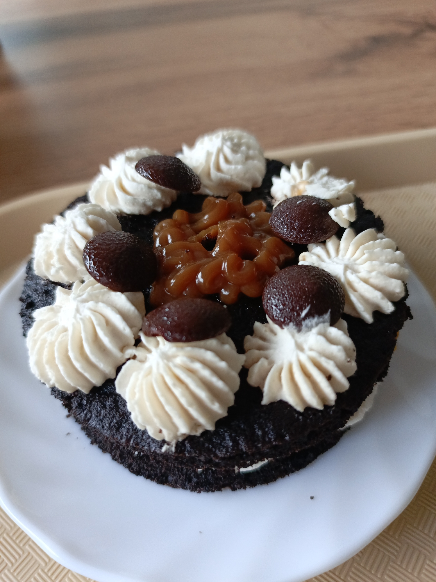 A dark-colored, circular dessert rests on a white, slightly rectangular plate with rounded edges. The dessert is topped with approximately twelve swirls of light-colored frosting, arranged around the perimeter and interior. Interspersed among the frosting swirls are seven small, dark brown, glossy, oval-shaped items, possibly dates or small fruits. At the center of the dessert, a cluster of translucent, amber-colored, viscous topping is present. The white plate sits on a light brown surface with a fine, woven texture. The background consists of a blurred, warm-toned wooden or laminate surface. No individuals are present. No identifiable features within the image indicate the city of Noviy, Russia.