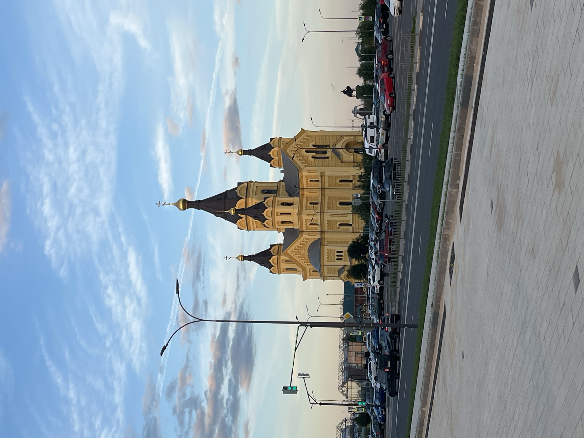 The image depicts a wide-angle view of the Alexander Nevsky Cathedral, a prominent Orthodox church located in Nizhny Novgorod, Russia. The cathedral, painted a pale yellow with dark domes and golden crosses, is the central subject of the photograph. It stands majestically against a backdrop of a soft, partly cloudy sky, with hues of blue and scattered white and pinkish clouds suggesting it is either dawn or dusk.
In the foreground, a paved walkway with large, rectangular grey tiles leads towards a street. This street is busy with traffic, showing a variety of cars parked and moving. The vehicles are a mix of colors and types, including sedans, SUVs, and vans. A traffic light with a green signal is visible on the left side of the image, indicating traffic flow. Several streetlights with curved arms are positioned along the road, some with multiple lamps.
Beyond the street and the parked cars, more buildings are visible, some with greenhouse-like structures, suggesting a commercial or industrial area adjacent to the cathedral. A large parking lot is filled with vehicles. Further in the background, a silhouette of a monument, possibly a statue on a horse, is visible against the sky. The overall environment is urban, with infrastructure like roads, streetlights, and buildings clearly defining the setting. There are no visible people in the image.