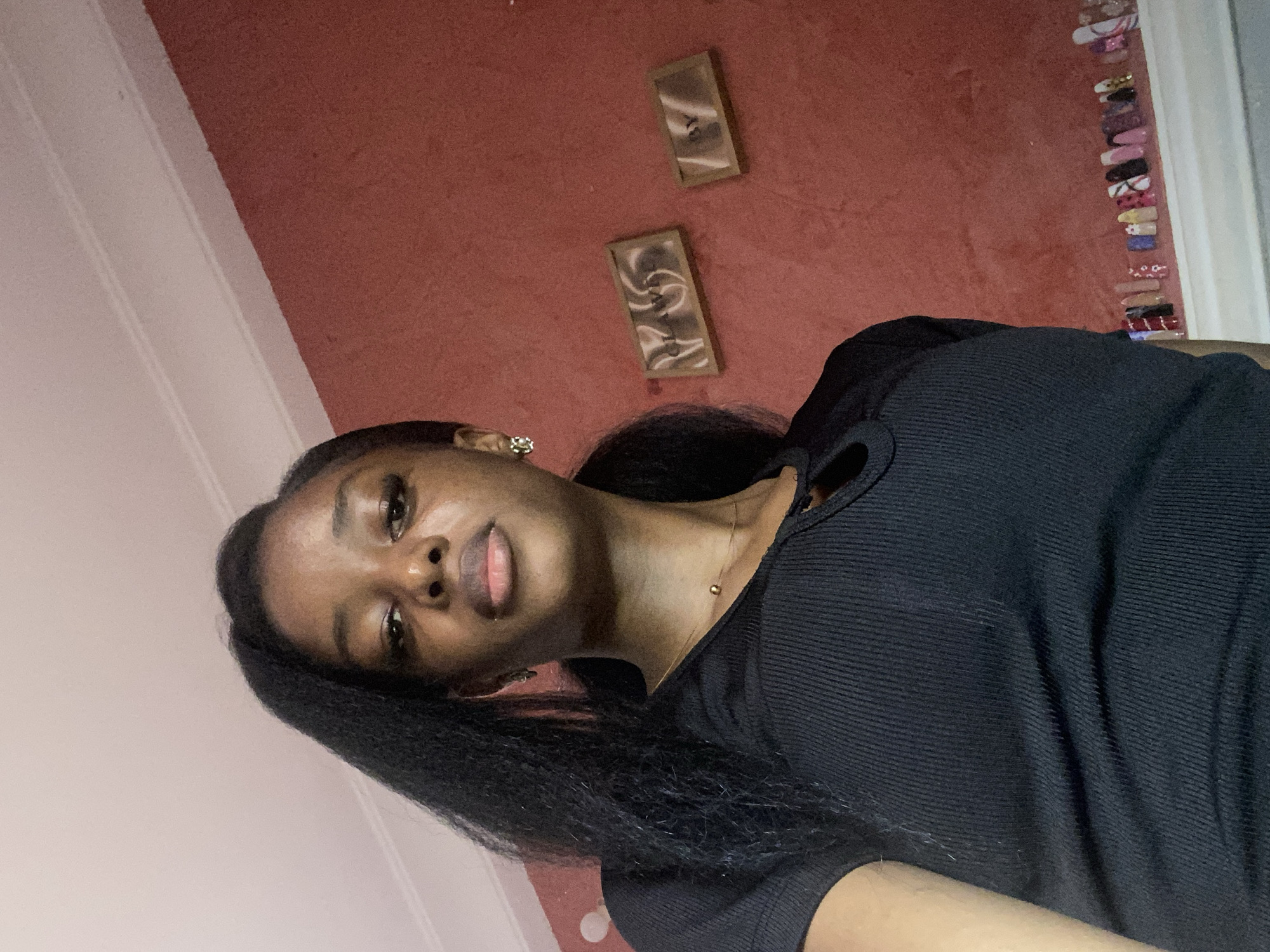 A young Black female is shown from a low angle in a close-up shot. She is wearing a black ribbed top with a keyhole neckline and a small gold pendant necklace. Her dark hair is styled in a long ponytail, with some strands falling over her shoulder. She has on earrings and what appears to be makeup. The background consists of a textured, coral-colored wall with two framed pictures hanging on it. To the right, a row of decorative artificial nails is displayed on a white ledge. The image's lighting suggests an indoor setting, potentially a residential room in Jos, Nigeria.