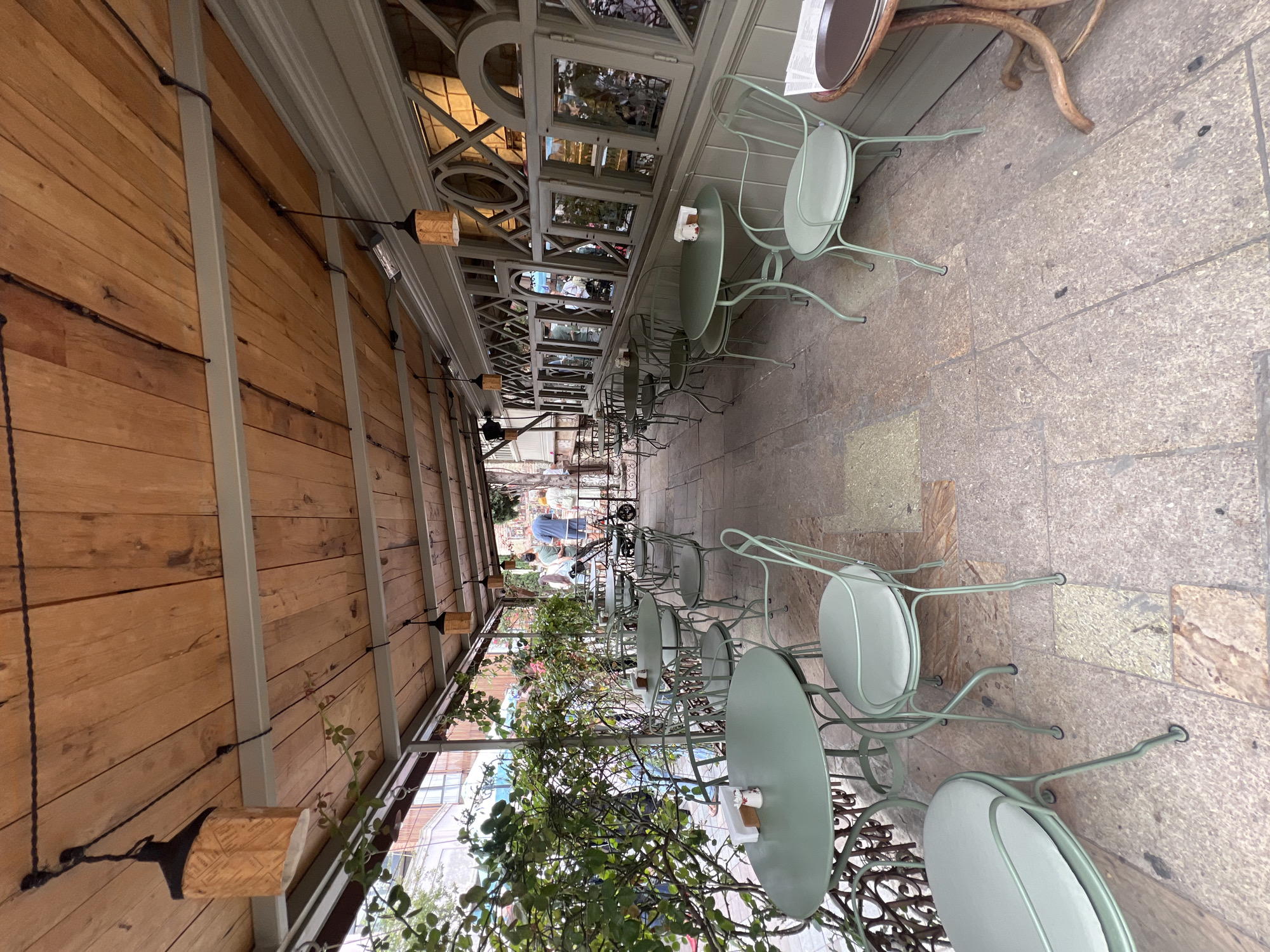 The image shows an outdoor seating area of a cafe in Tbilisi, Georgia, with a covered patio. The patio roof is made of wooden planks, and pendant lights with wooden accents hang from it. Green metal tables and chairs are arranged on a stone tiled floor. Lush green foliage from a climbing plant covers the left side of the frame, obscuring some of the tables and chairs. The building facade to the right features a series of windows with decorative lattice work. In the background, several people are visible, appearing to be patrons of the cafe. One person is standing near a bicycle or scooter. The overall atmosphere suggests a casual, daytime setting.