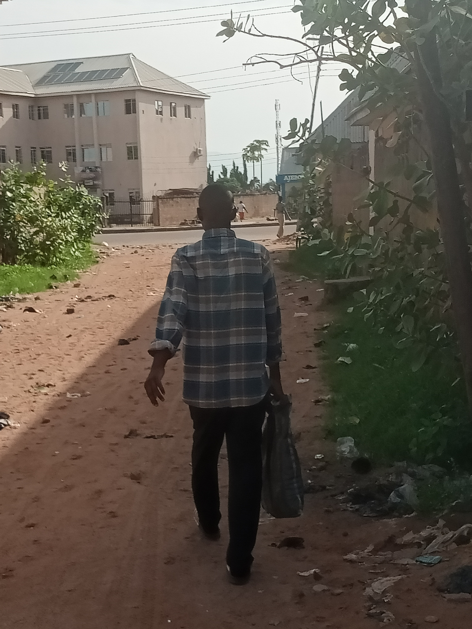 A person with dark skin and a shaved head walks away from the camera on a dirt path lined with grass and debris. They are wearing a blue and white checkered long-sleeved shirt and black pants, carrying a grey bag in their right hand. In the background, a multi-story building with solar panels on its roof is visible, along with some trees and a cell tower. The scene appears to be in a city environment, possibly Jalingo, Nigeria. The lighting suggests it is daytime, with the sun casting shadows on the path. The overall atmosphere is somewhat unkempt, with visible litter on the ground.