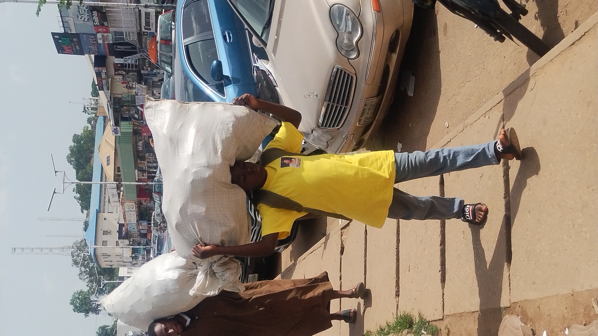 The image shows two individuals carrying large sacks on their heads in an urban area. The person in the foreground wears a yellow t-shirt, blue jeans, and sandals. He is carrying a white sack over his head and has a bag slung over his shoulder. A second person, partially obscured, is carrying a similar sack. Behind them is a parked white Mercedes-Benz sedan with Nigerian license plate. Further back, there are several other vehicles and commercial buildings visible, including billboards advertising various businesses. The presence of billboards and commercial buildings suggests the location is within a city, specifically, Jos, Nigeria. The ground is paved and there is some vegetation growing nearby. The scene appears to be a street or sidewalk with commercial activity.
