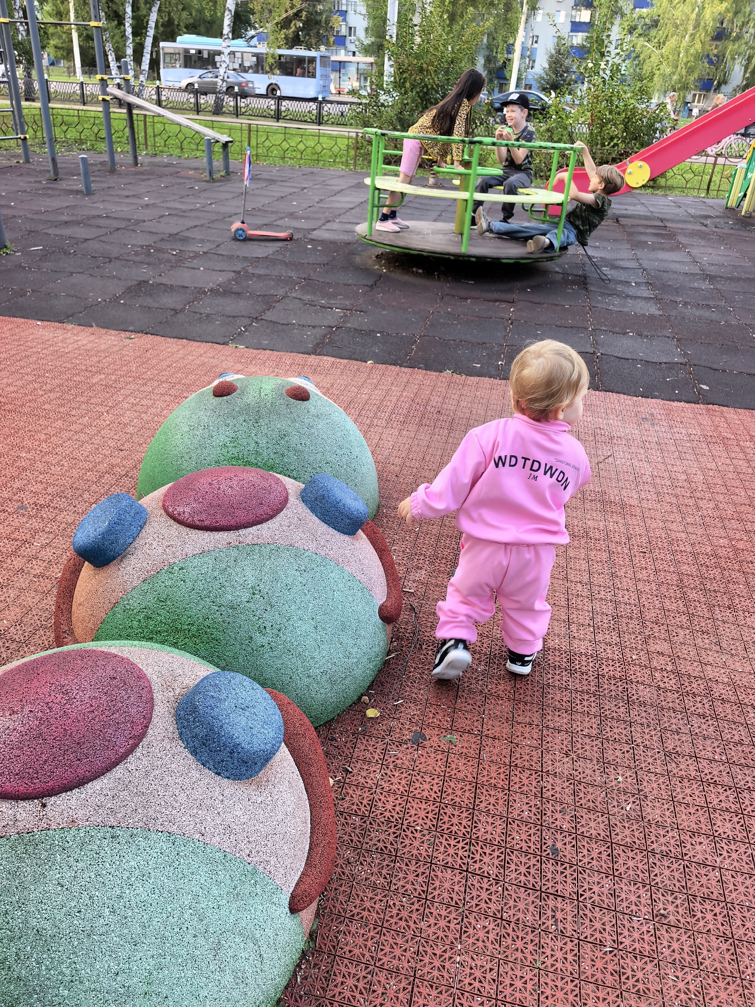 This image captures a playground scene likely located in the city of Sterlitamak, Russia. In the foreground, a young child, seen from behind, is wearing a pink tracksuit and black and white sneakers, walking away from the camera on a red, textured playground surface. To the left, large, round, colorful climbing structures with red, green, and pink sections and blue accents are visible. In the middle ground, a group of older children are interacting on a merry-go-round and a red slide. One child is on the merry-go-round, another is reaching for the slide, and a third is seated on the merry-go-round. A scooter is parked on the black rubberized playground surface. In the background, a blue bus and other vehicles are visible, suggesting the playground is situated in an urban or suburban environment with passing traffic. Trees and apartment buildings line the perimeter of the playground area.