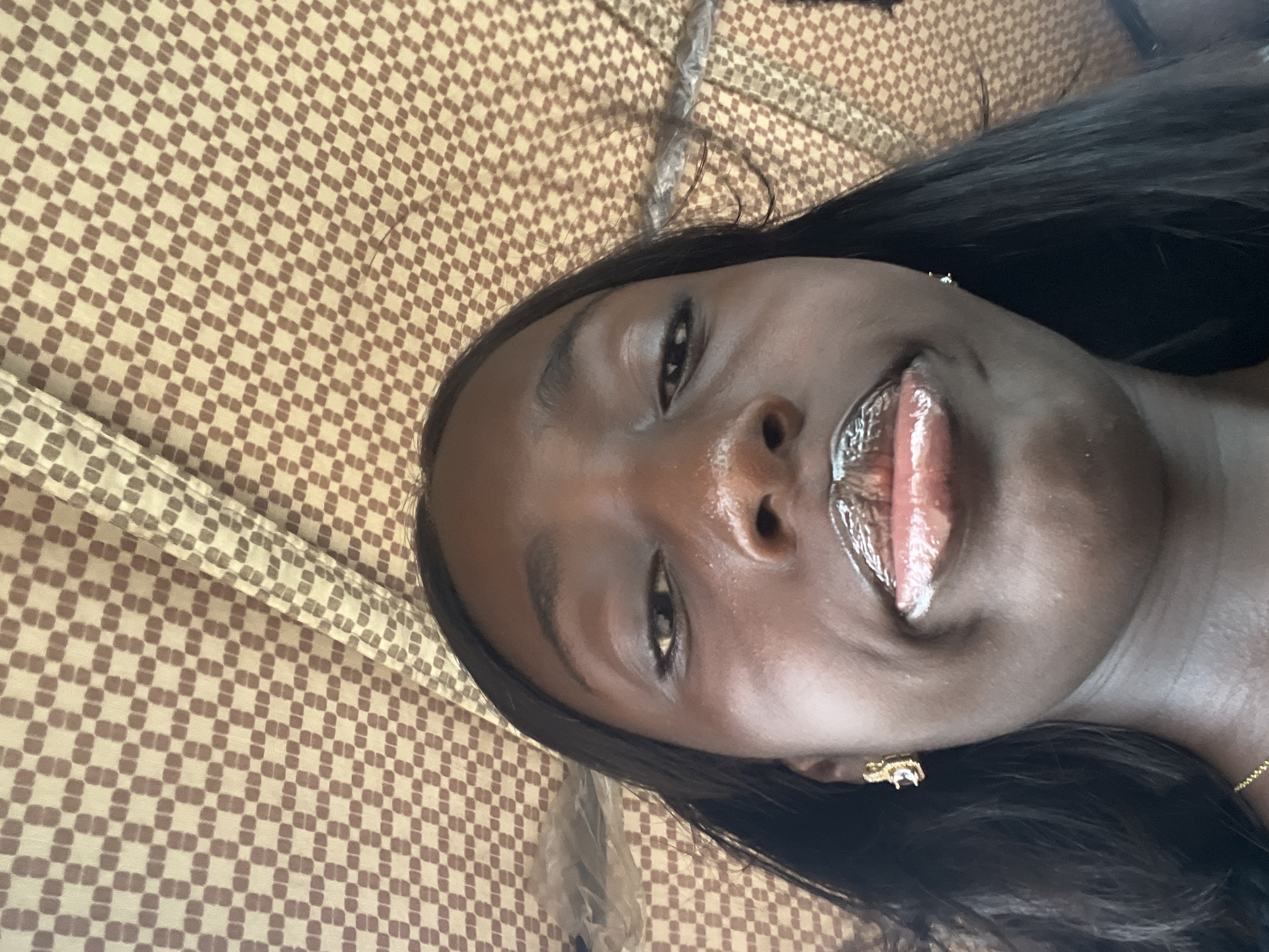 A medium shot of a person with dark brown skin and straight, black hair, set against a background of a light brown and white checkered pattern, possibly a fabric. The person is wearing what appears to be gold earrings and lip gloss. It is difficult to determine anything definitive about the location in Galadimawa Village, Nigeria based on this image.