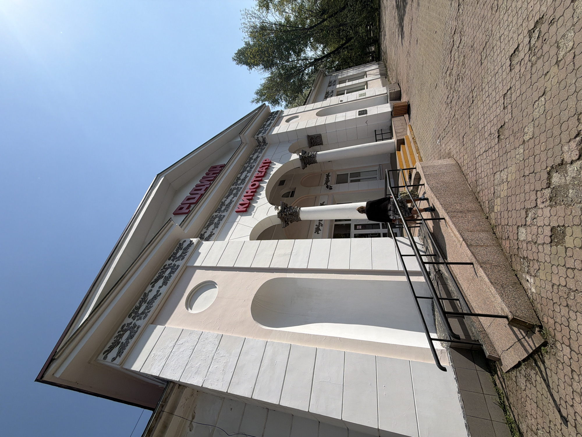 The image shows a building in Birobidzhan, Russia. The building's facade is white with decorative elements, including red lettering that reads "RODINA KINOTEATR" (Homeland Cinema). The building has a classical architectural style with columns and arches. A person is standing near the entrance of the building, which has a black metal railing. The sidewalk in front of the building is paved with brown interlocking pavers. The sky is blue, and a tree is visible above the building.