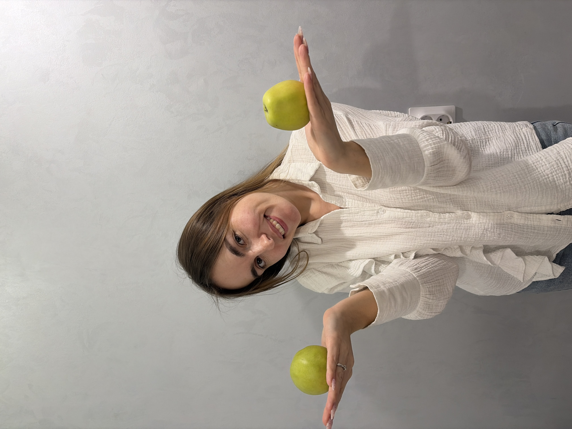 A woman is holding two green apples, one in each hand, while smiling at the camera. She's wearing a white, long-sleeved shirt. A white electrical outlet is visible on the wall behind her. The background is a gray, textured wall. The location is unknown, and there is no evidence to suggest it is in Neftekamsk, Russia.
