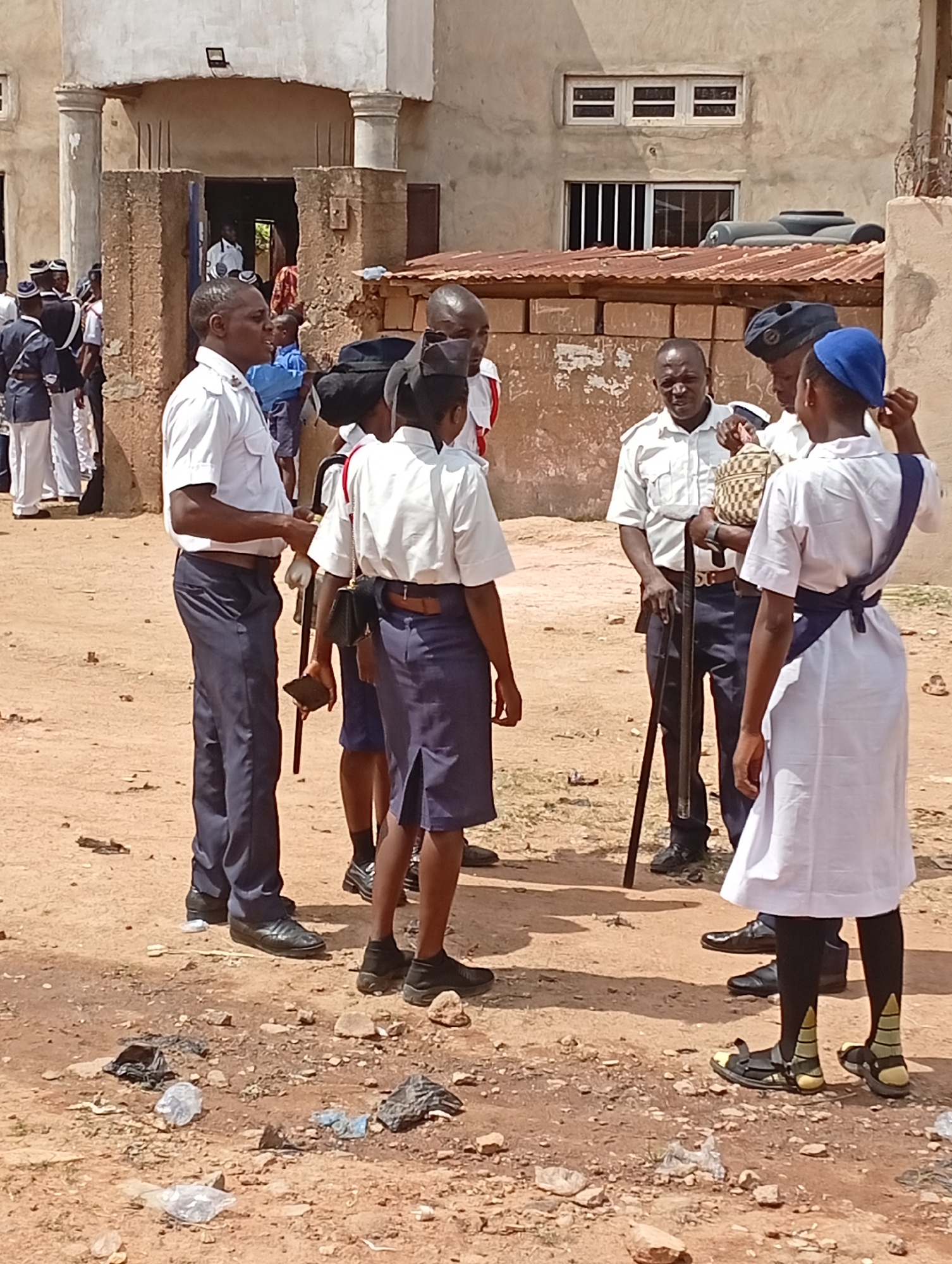 The image depicts a group of uniformed individuals in an outdoor setting. Several people wear white shirts and dark trousers or skirts. One person has a blue head covering and a white dress with a dark sash. Some individuals hold long, dark-colored sticks. The ground is dirt or sand, with scattered trash. Buildings appear in the background. The location is Unknown, Unknown.