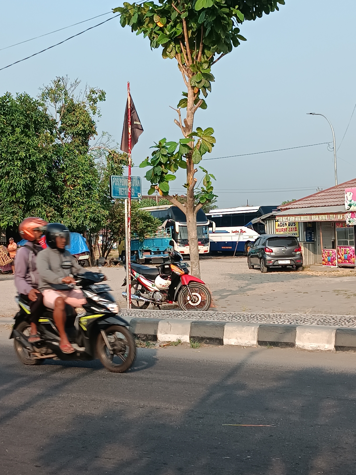 In Imogiri, Indonesia, an outdoor scene depicts a street with a paved road, a pedestrian walkway, and a parking area. A motorcycle with two occupants, both wearing helmets, is the primary focus in the foreground, moving from left to right. The passenger wears an orange helmet and a purple shirt, while the driver wears a black helmet and a grey long-sleeved shirt with pink shorts. Behind them, a red and black motorcycle is parked. In the background, a light blue bus and a dark grey car are visible. A small building with a brown roof, possibly a kiosk or shop, is situated in the middle ground. A sign above the building reads "AGEN BUS MURMT JAYA." Further back, more trees and a blue tent are present. A black flag with red writing flutters on a pole near a sign that says "POS PEMUNGUTAN RETRIBUSI." The sky is clear and blue, suggesting a sunny day.