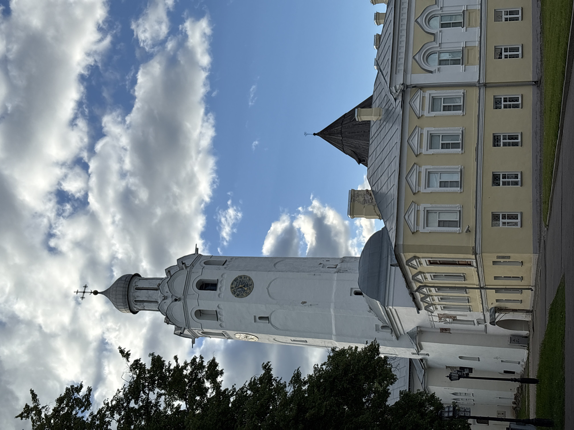 The image depicts a vertical view of buildings in Veliky Novgorod, Russia, beneath a sky with puffy white clouds and blue sky. A tall, white tower with a clock on its facade and a dome-shaped top with a cross stands prominently. Beside it is a yellow building with multiple rows of windows and a grey roof. Trees are at the bottom of the image, partially obscuring the lower sections of the buildings.