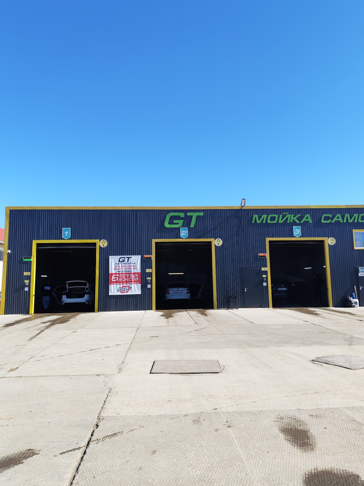 The image shows the exterior of a "GT" car wash building in Chaikovsky, Russia. The building has a dark gray corrugated metal facade with yellow trim. There are three bays visible, numbered 1, 2, and 3. Cars are inside bays 1 and 2, undergoing washing or detailing services. There is a sign advertising the car wash as a place to save time and stating that it offers "6 heated boxes." A person stands in front of the left most bay. The foreground shows a concrete apron, marked with water stains. A clear blue sky is visible above the building.