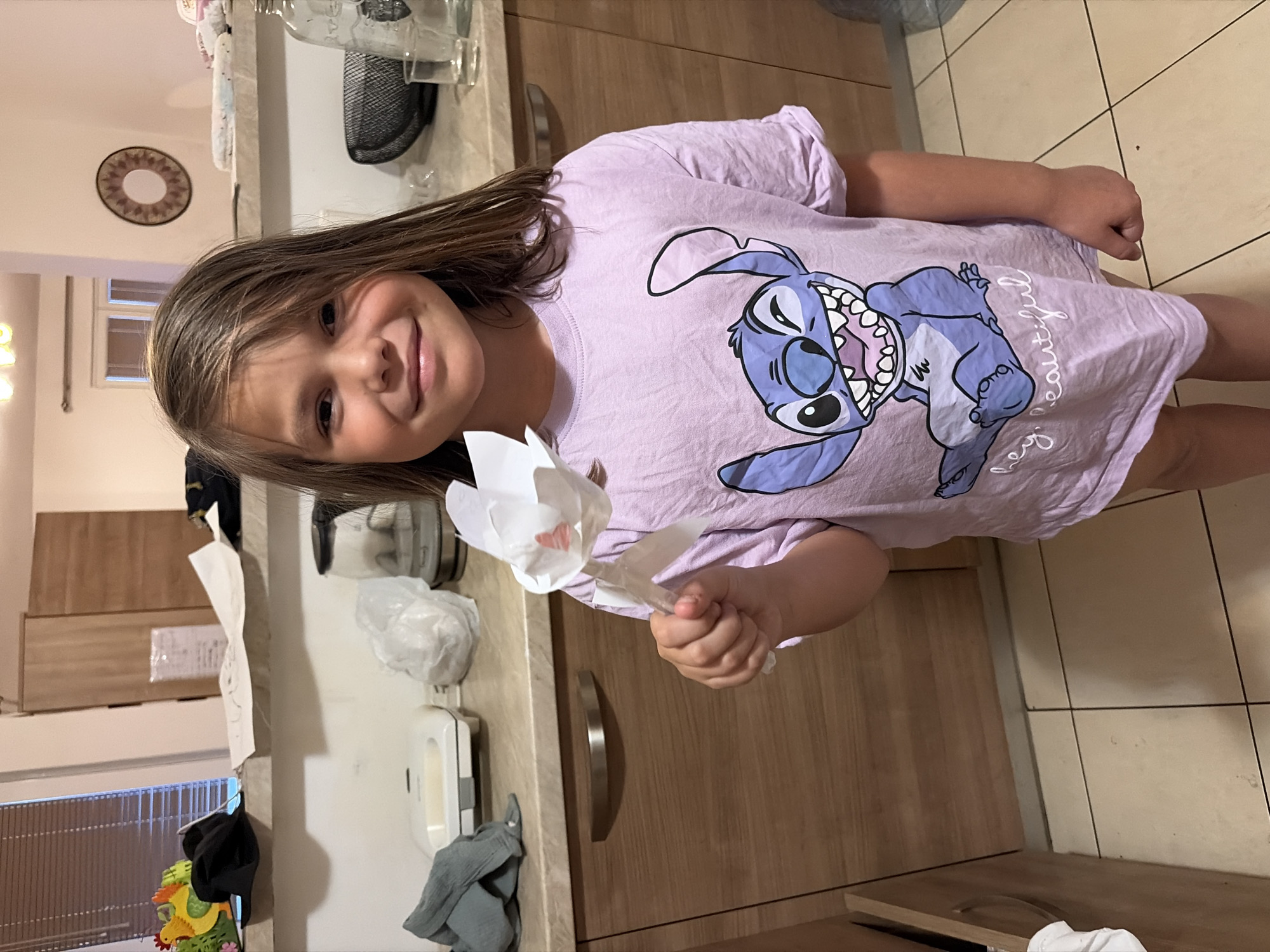 A girl is holding a paper flower. She is wearing a purple shirt with a cartoon character on it. She is standing in a kitchen with wooden cabinets and a tiled floor. The location is in Budva, Montenegro.